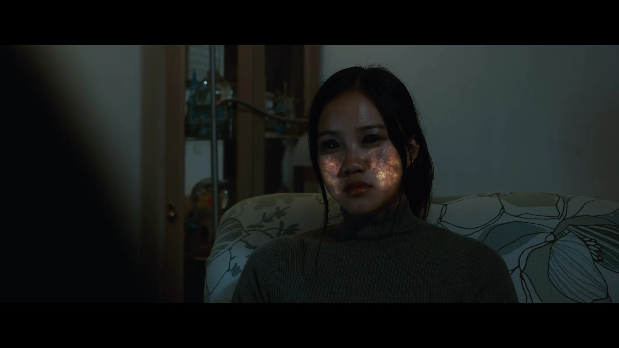 A woman sitting on a patterned sofa in a dimly lit room, with her face illuminated by a red radiation intertwined in her skin.