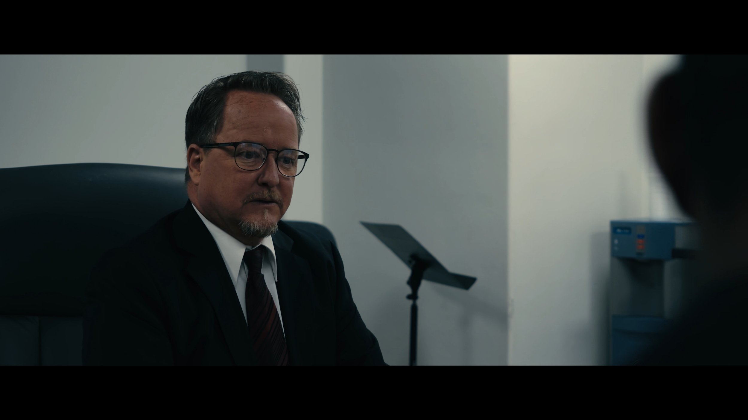 A man with glasses and a beard wearing a black suit and tie sitting in an office.