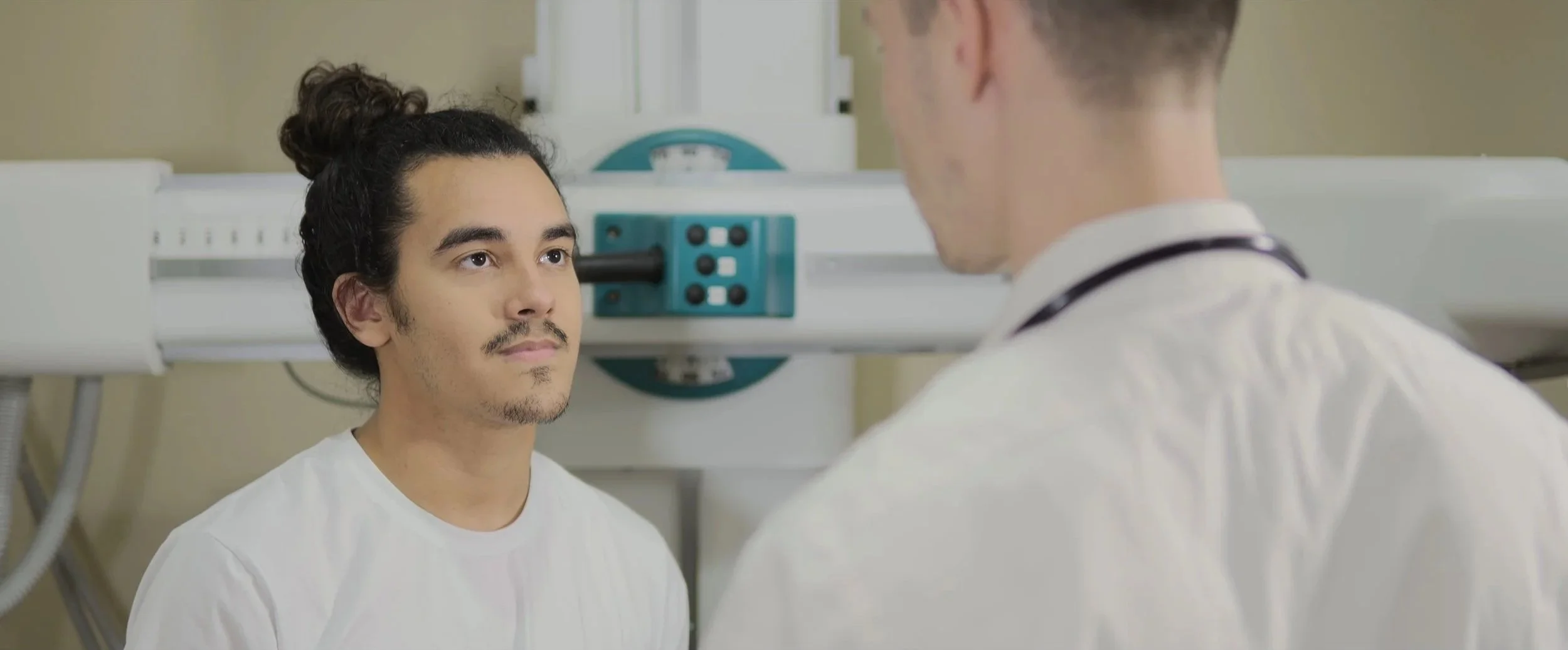A male patient with long dark hair, facial hair, and a white shirt sitting in a hospital or medical setting, facing a doctor wearing a white coat. The doctor has a stethoscope around his neck, and both are engaged in conversation. Medical equipment is visible in the background.