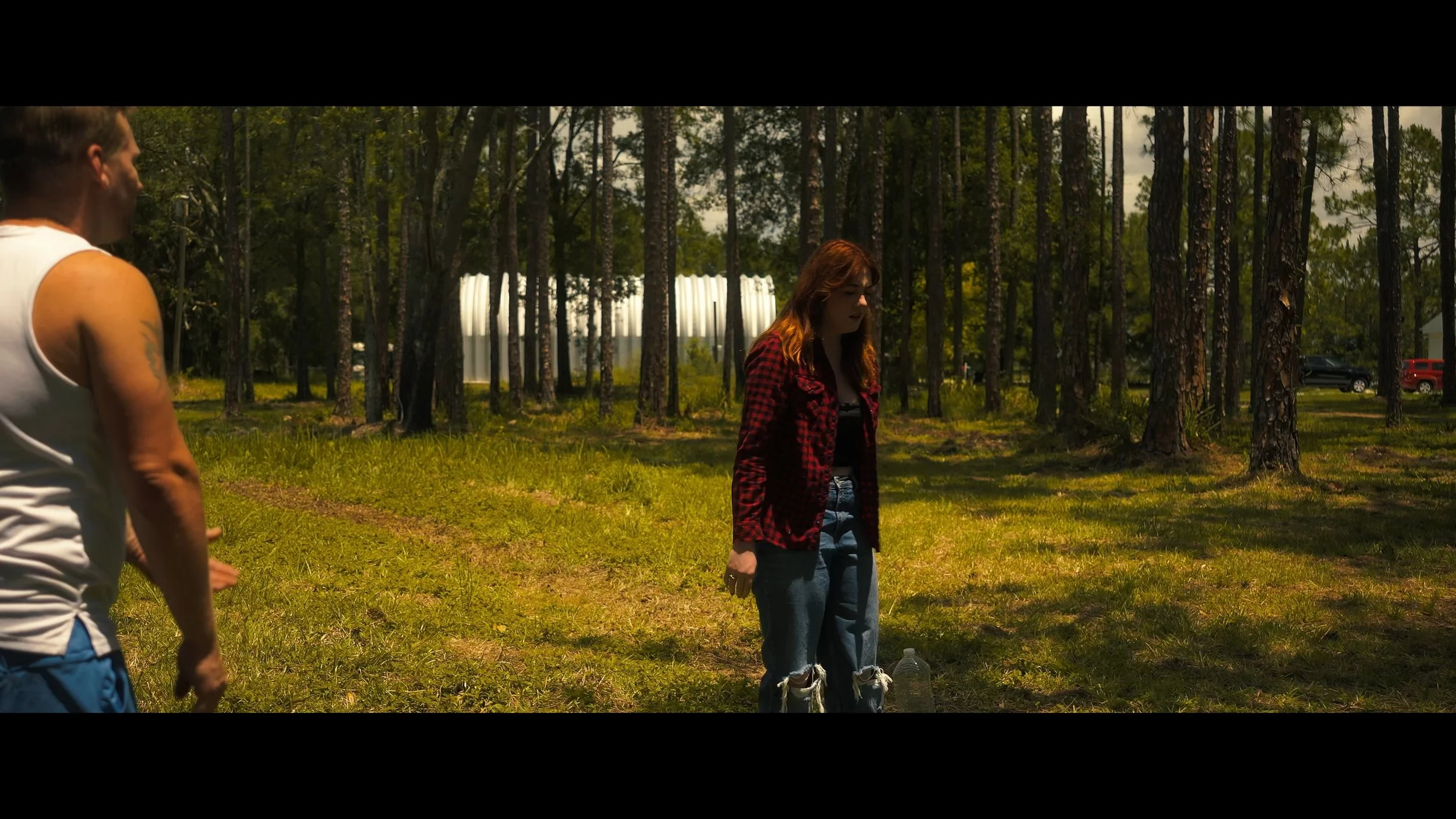 Three people outdoors in a forested area during daytime, one man on the left wearing a sleeveless shirt, a woman in the center wearing a red plaid shirt, and part of another person is visible on the right, with a background of tall trees and parked c
