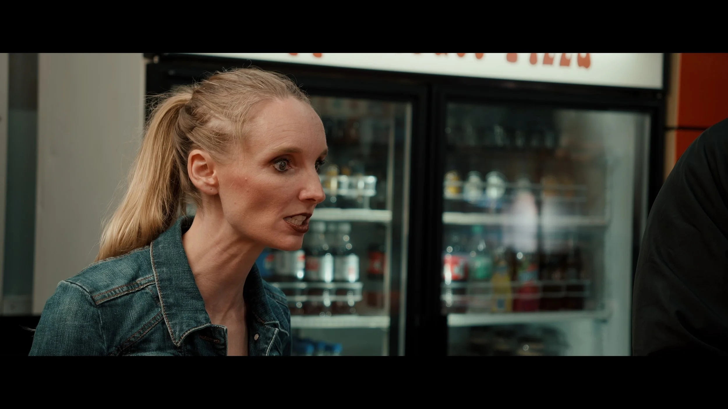 A woman with blonde hair tied back, wearing a denim jacket, appears to be speaking intensely in a store or convenience shop, with refrigerated beverage units in the background.