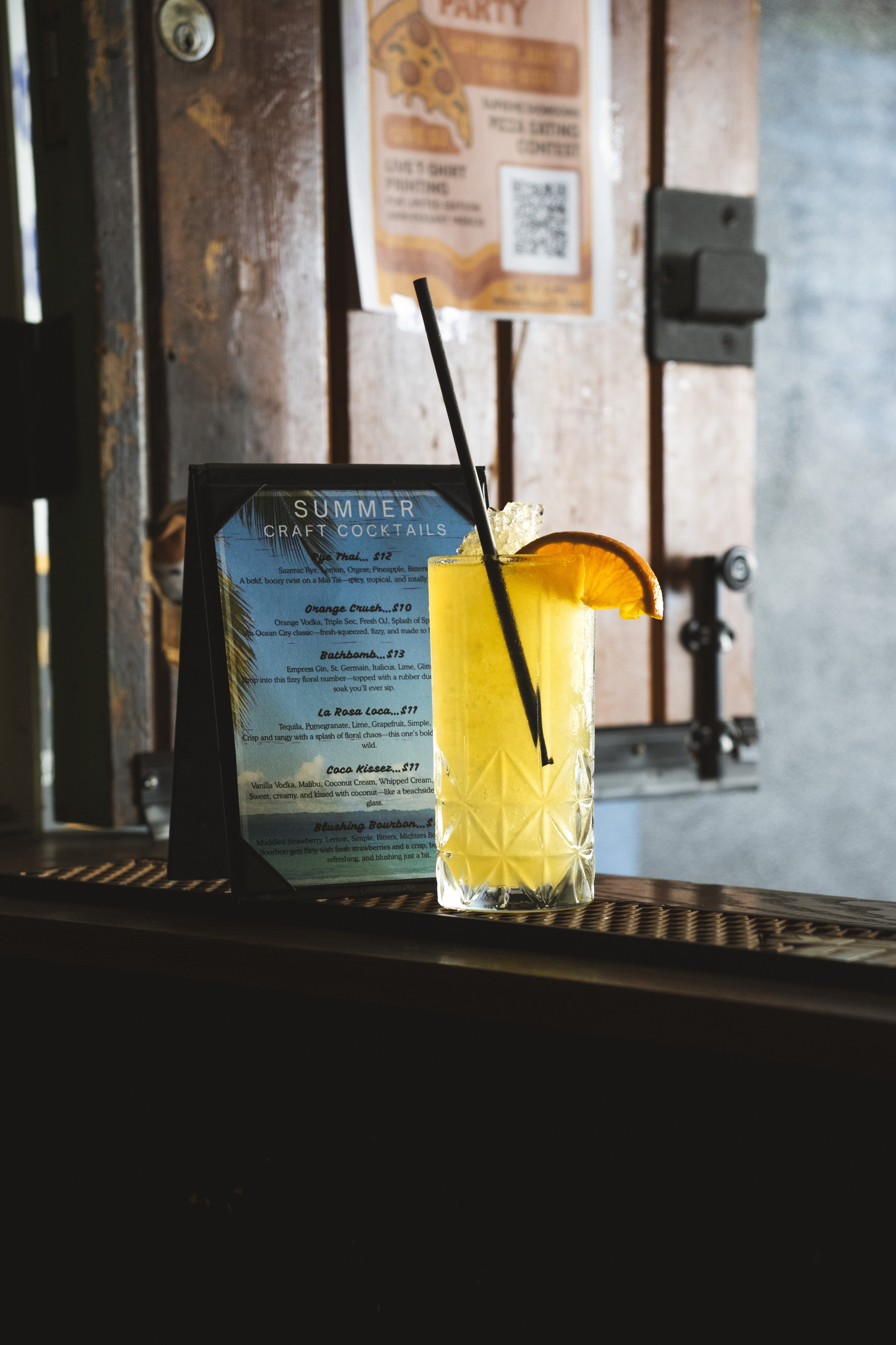 A tall, yellow cocktail with an orange slice garnishing the rim, a black straw, placed on a bar counter, with a menu and wooden wall in the background.