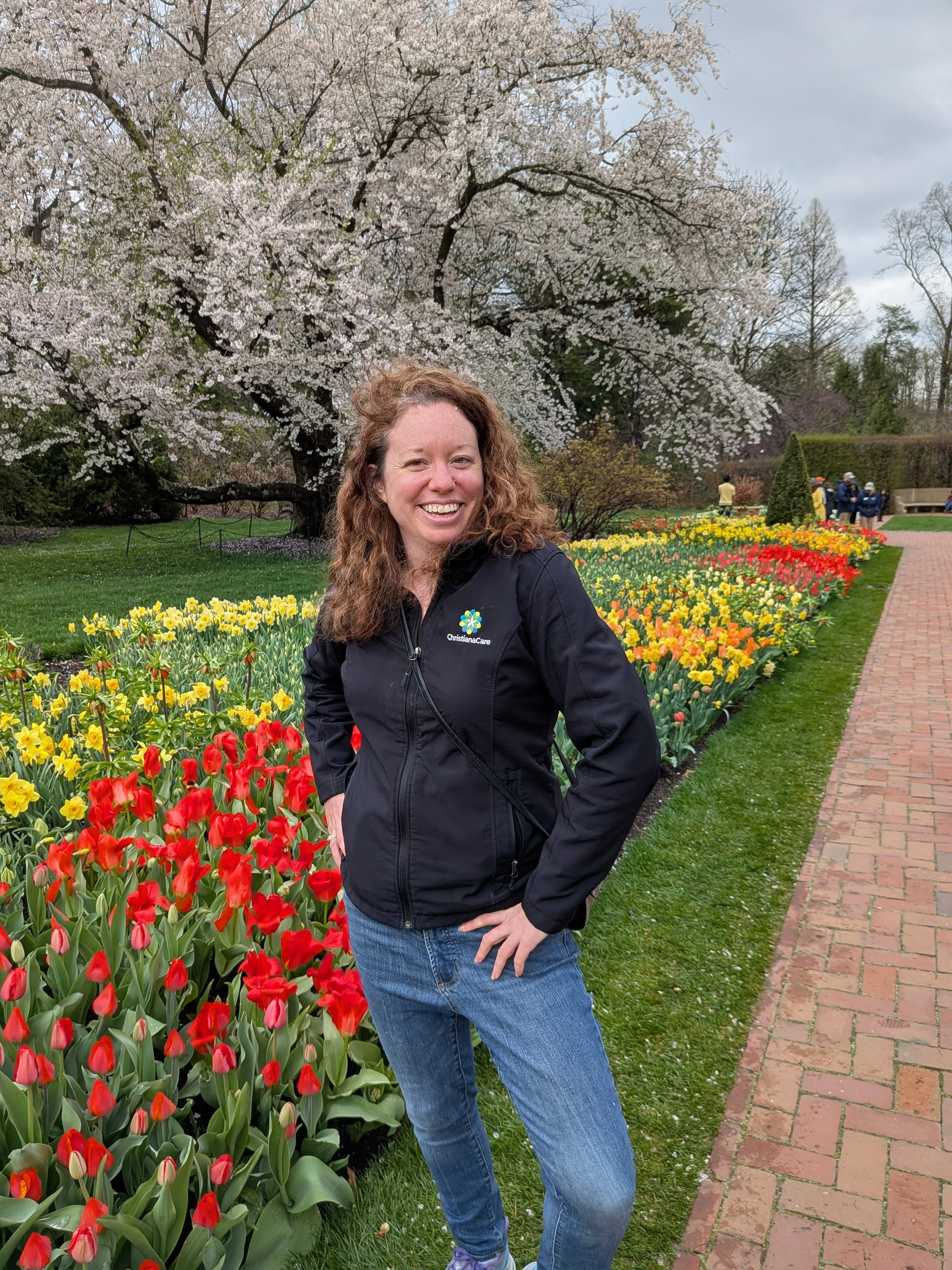 A woman with curly brown hair smiling and standing in a flower garden with colorful tulips, a large blooming cherry blossom tree, and a pathway with other people in the background.