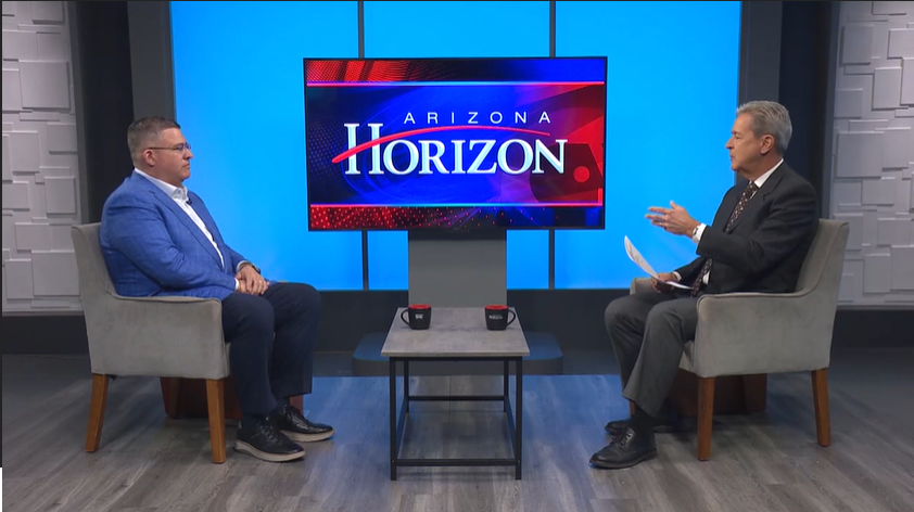 A televised interview with two men seated on stage in front of a large screen displaying 'Arizona Horizon'. One man is wearing a blue blazer and glasses, the other in a black suit, engaging in conversation.