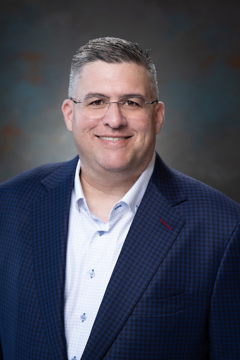 Headshot of a man wearing glasses, a white dress shirt, and a dark blue blazer, smiling at the camera.
