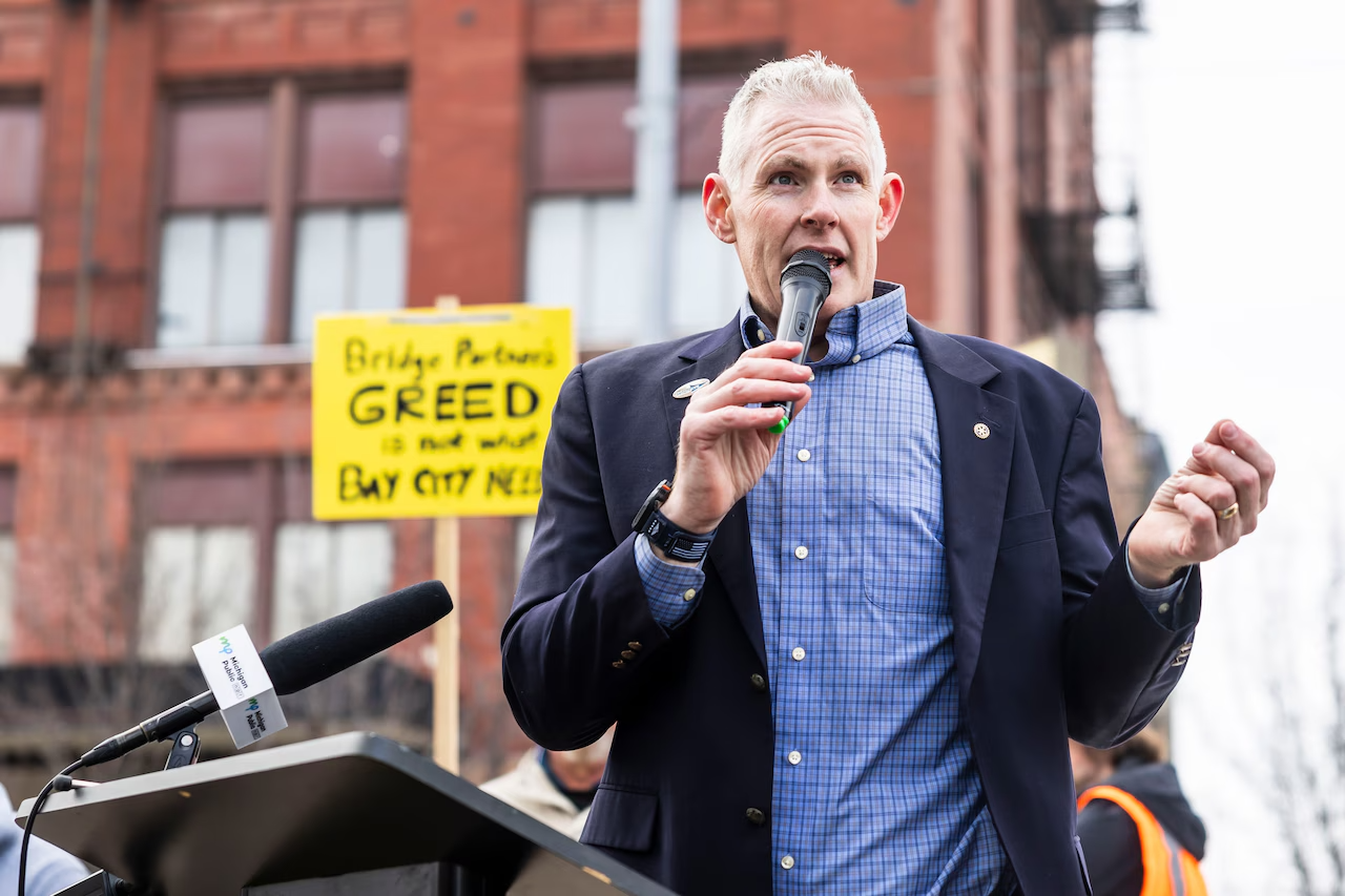 Chris Girard in a blue shirt and blazer speaking into a microphone during a protest or rally. In the background, there is a yellow sign that reads 'Bridge Polluters GREED is not okay Bay City Needs'.