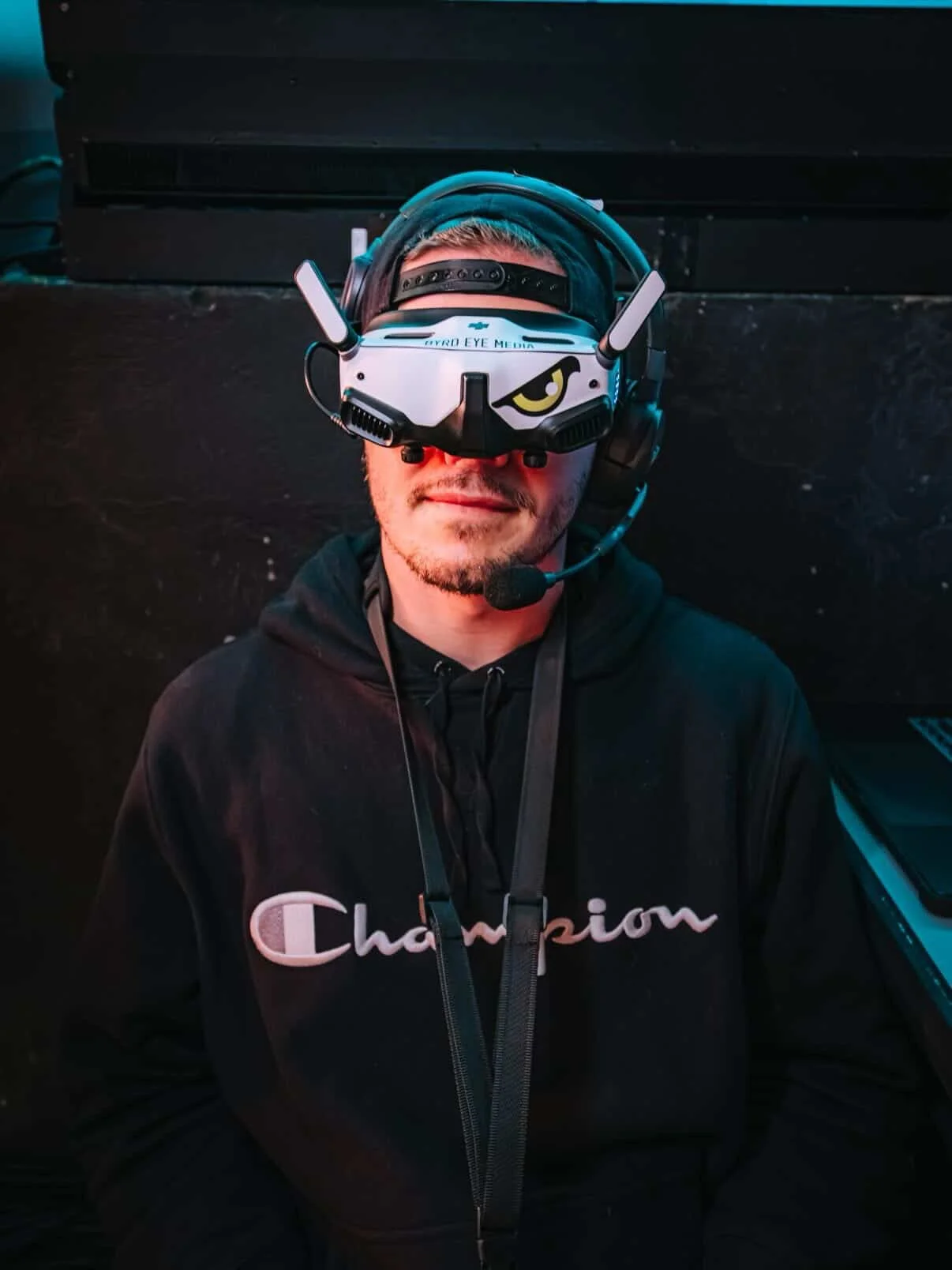 A man wearing a virtual reality headset with a helmet and microphone, dressed in a black hoodie with the word 'Champion' on it, sitting against a dark wall.