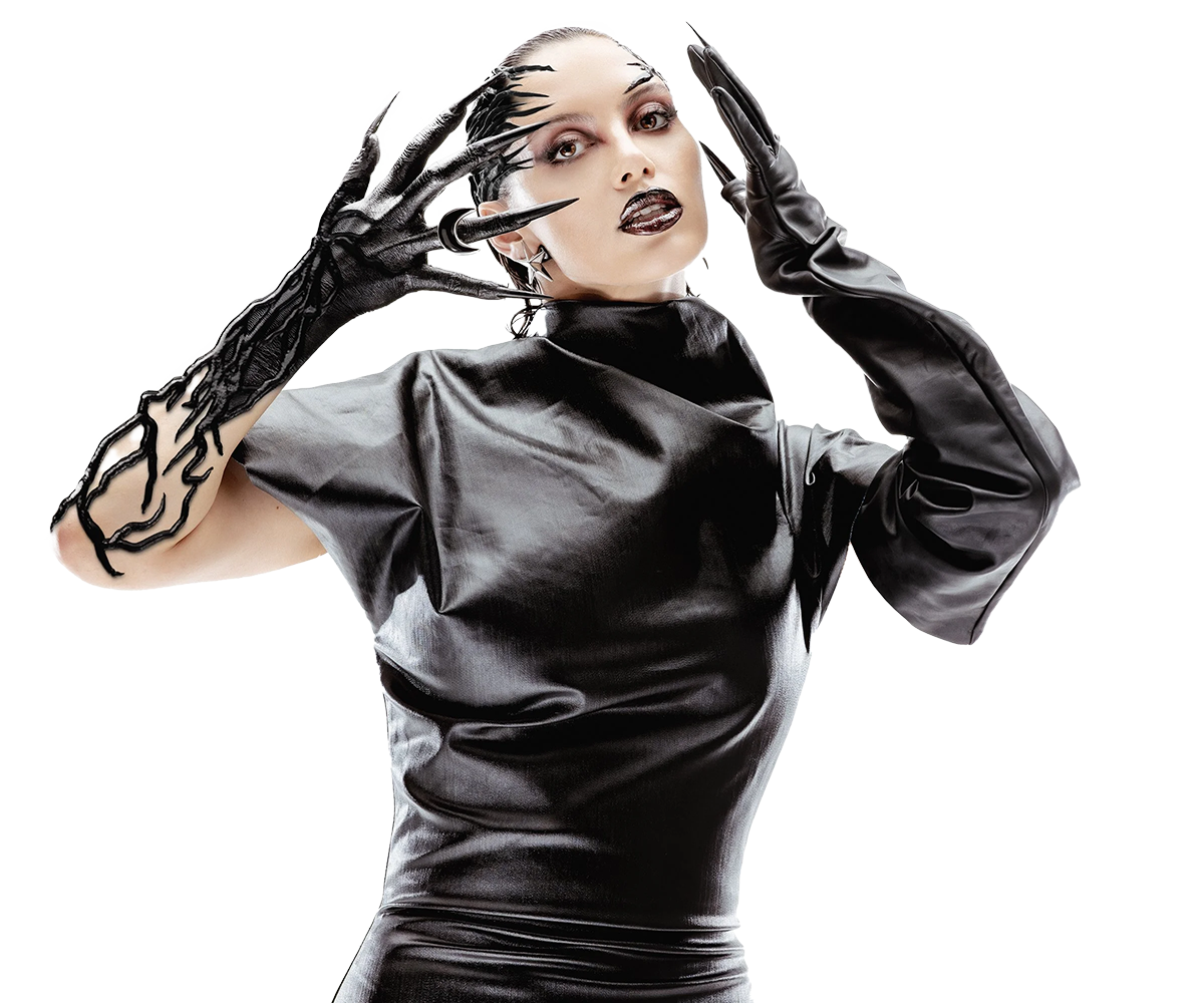 A woman with pale skin wearing a black shiny dress and long black gloves. She has bold makeup with dark lipstick and eye makeup, and her hair is styled back. She is holding her head with one hand and has her fingers near her face, striking a dramatic pose against a black background.
