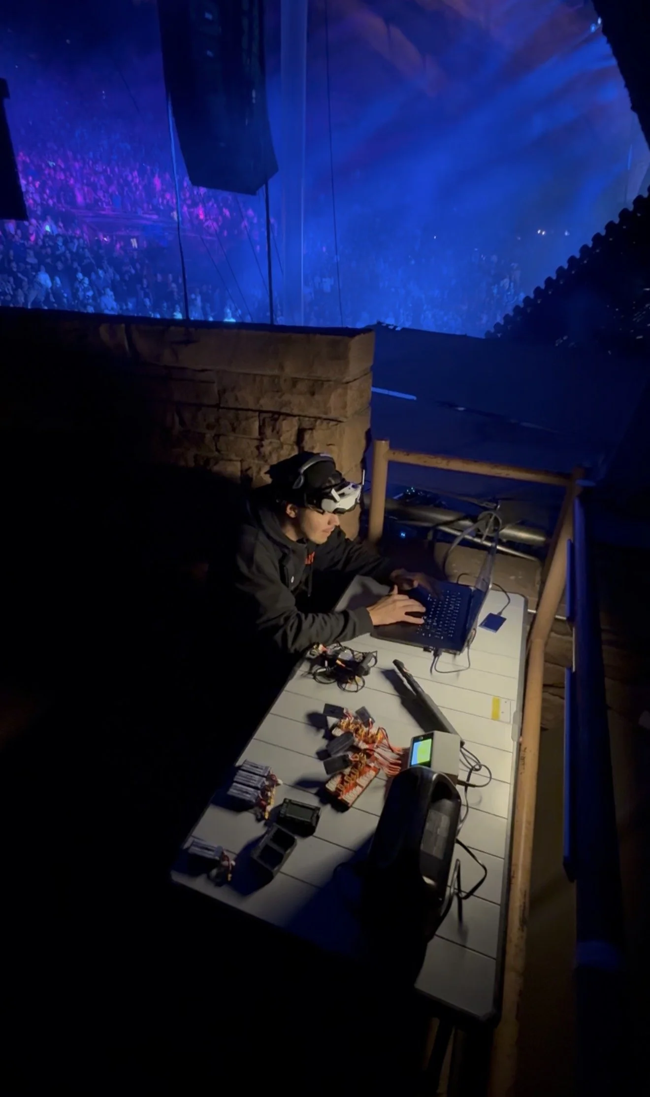 Person wearing a virtual reality headset working on a laptop at a table with electronic components and wires, in a dark room with a large audience projected on the wall in the background.