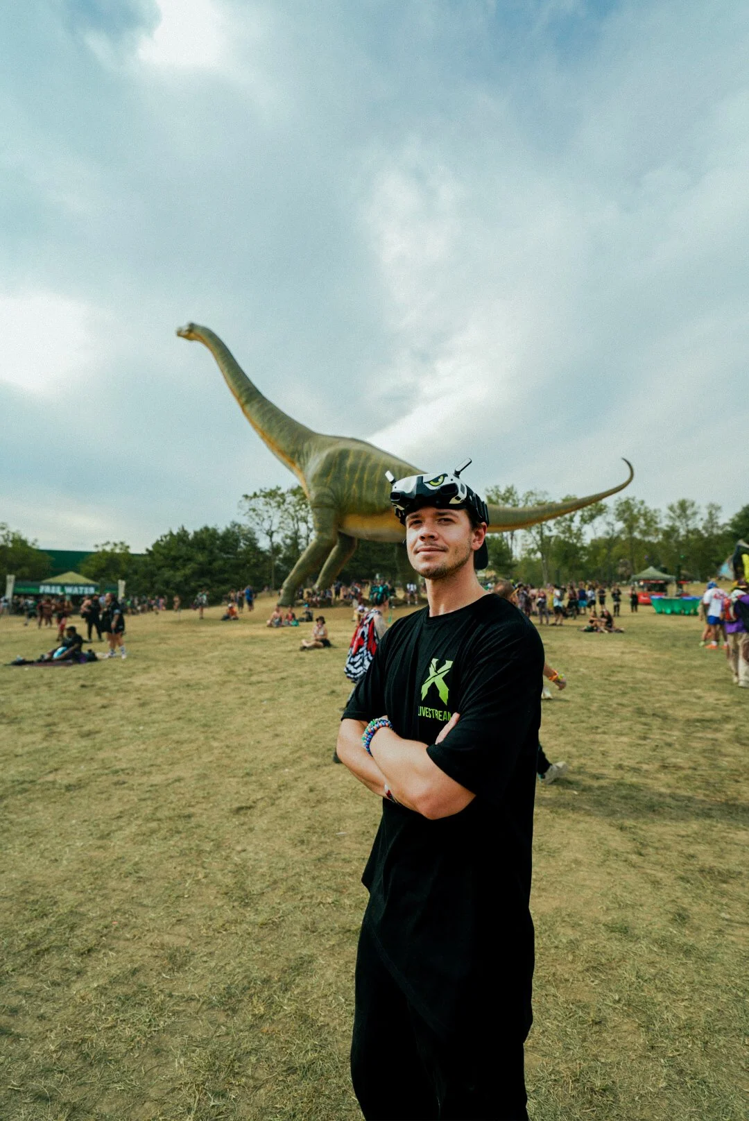 A young man wearing a black t-shirt and a drone pilot headband stands with arms crossed in a park with a large dinosaur sculpture and many people in the background.