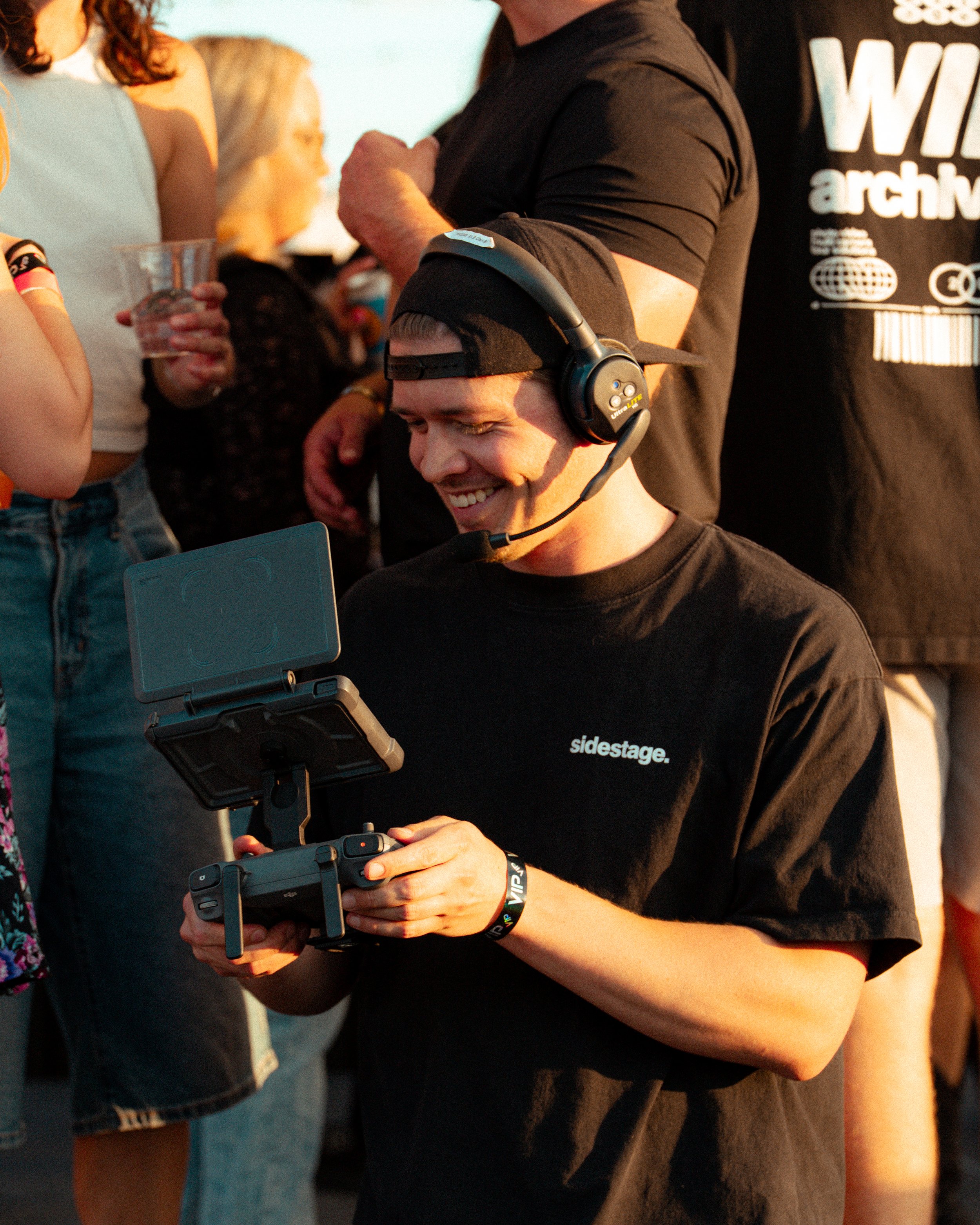 A person wearing a black t-shirt with white text and a black baseball cap with a headset, smiling while operating drone controls. Other people are holding drinks and socializing in the background.