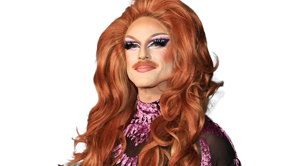 A transgender drag queen with long red hair styled in loose waves, wearing bold makeup with purple eyeshadow, thick eyeliner, and pink lipstick, dressed in a purple patterned top.