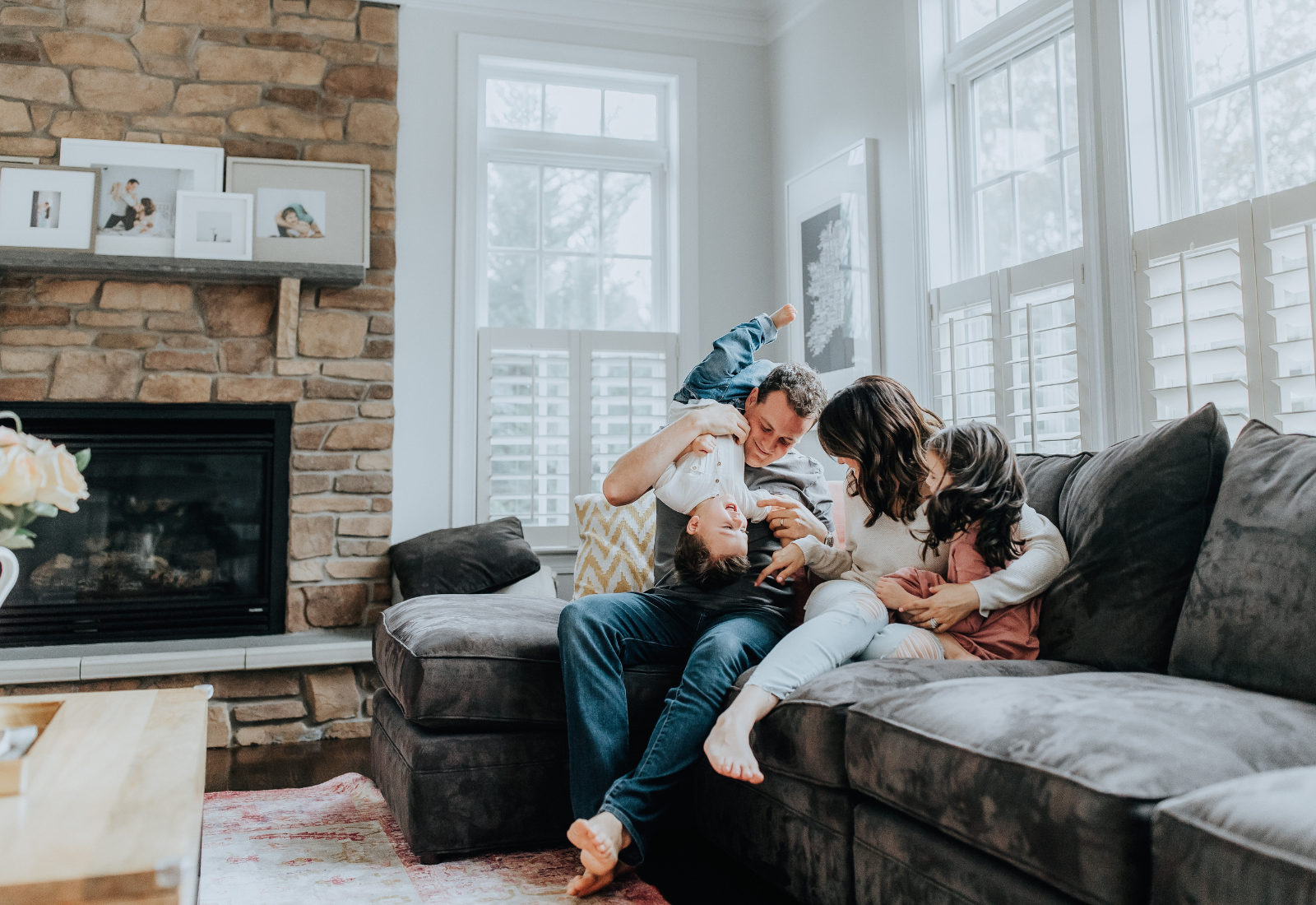 A family of four playing and laughing together on a gray couch in a bright living room with large windows and a fireplace.