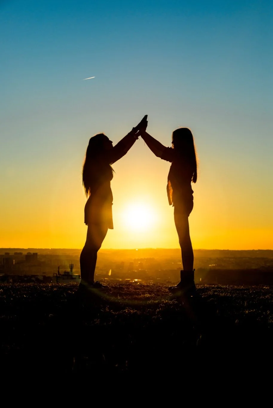 Silhouettes of two girls high-fiving against a sunset background, with a cityscape in the distance.