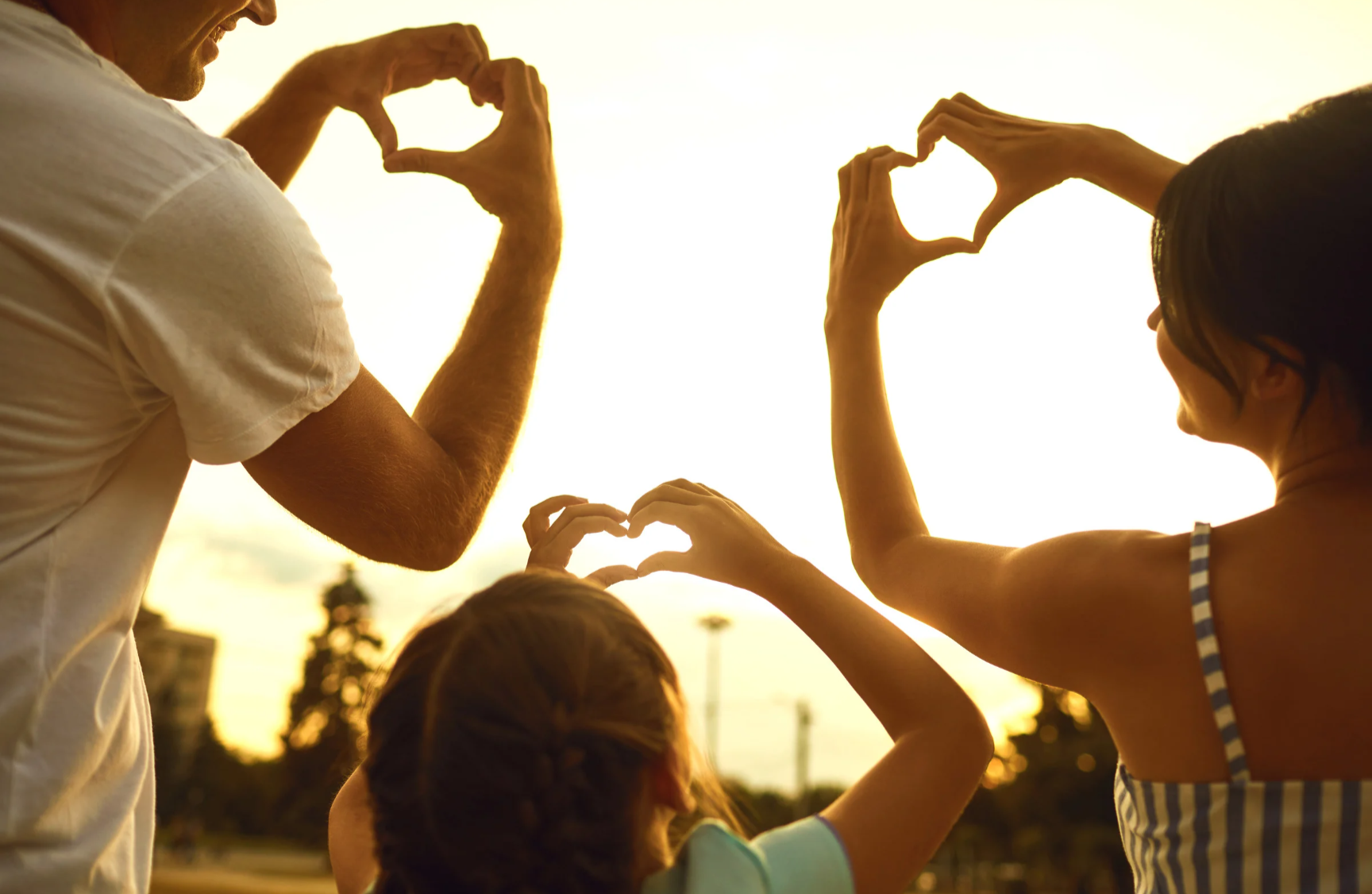People outdoors at sunset forming heart shapes with their hands, including children and adults.