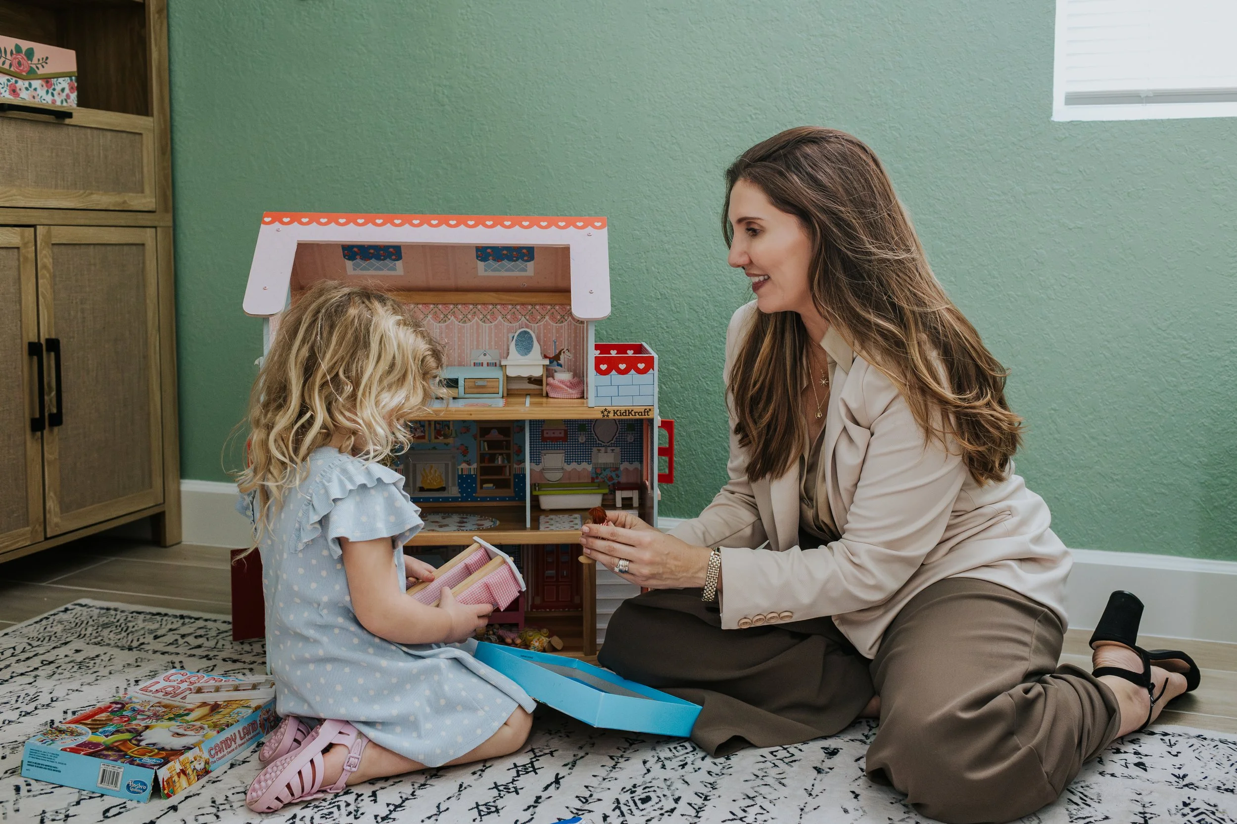 A woman and a young girl playing with a dollhouse inside a room. The woman is kneeling on the floor, smiling at the girl, who is sitting on the rug. The room has a green wall and wooden furniture. The girl is wearing a light blue dress with ruffles, and the woman is dressed in a beige blazer and dark pants.