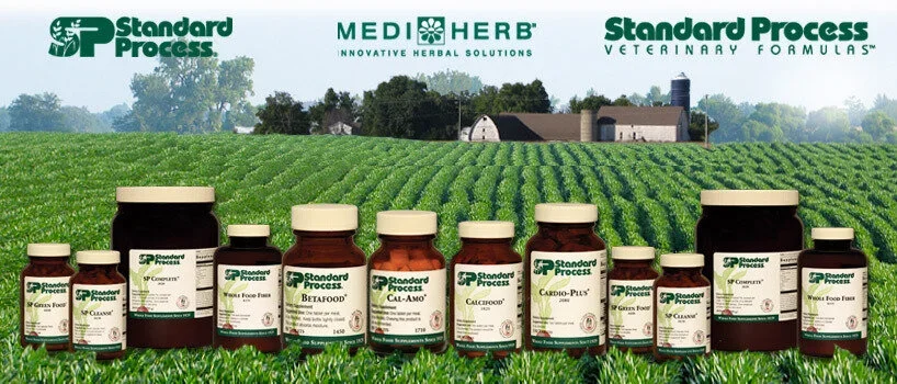 Multiple bottles of Standard Process and MediHerb herbal supplements arranged on grass in front of a farm and green fields under a cloudy sky.