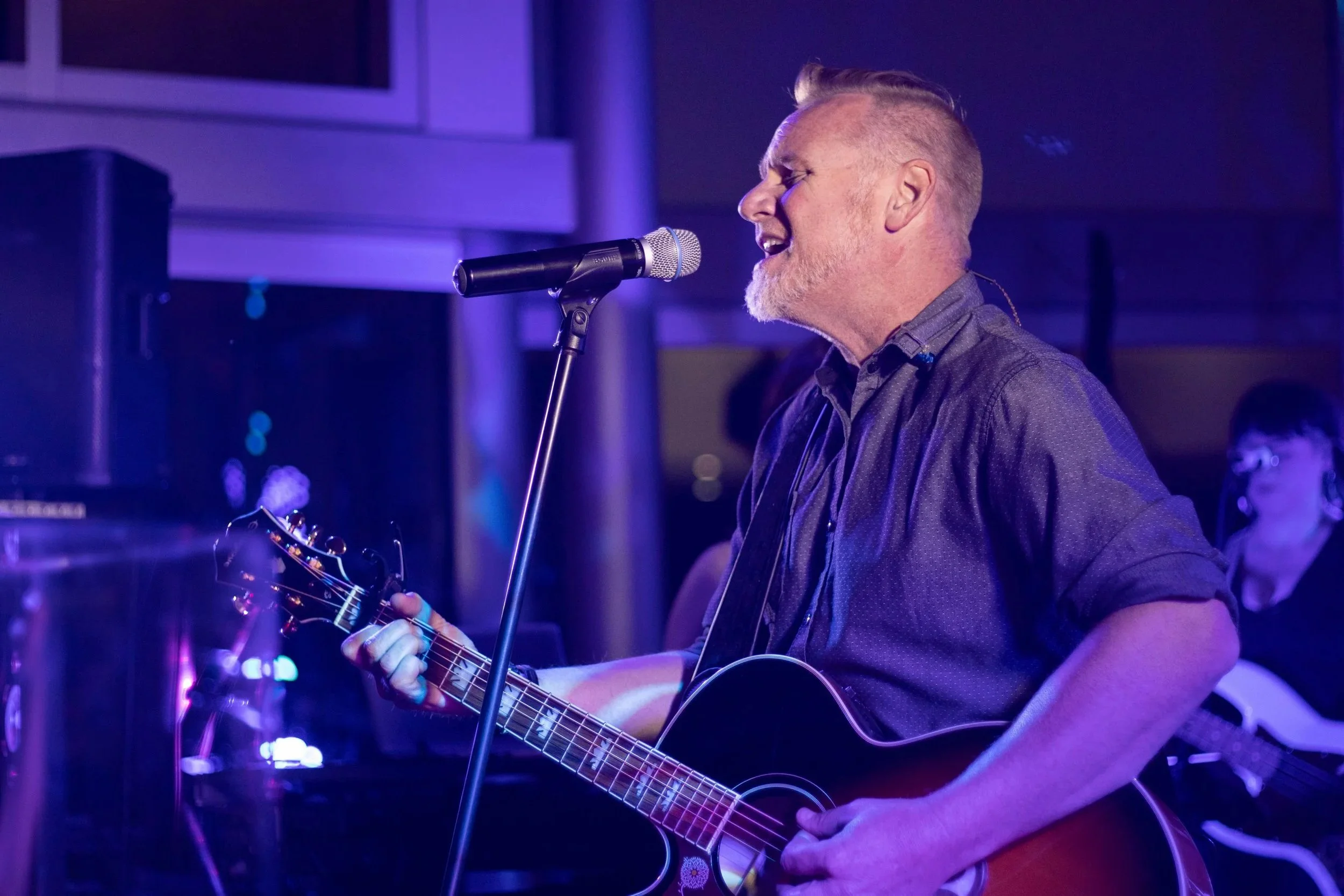 A man with a beard and short blonde hair singing into a microphone while playing an acoustic guitar in a dimly lit room with purple lighting.