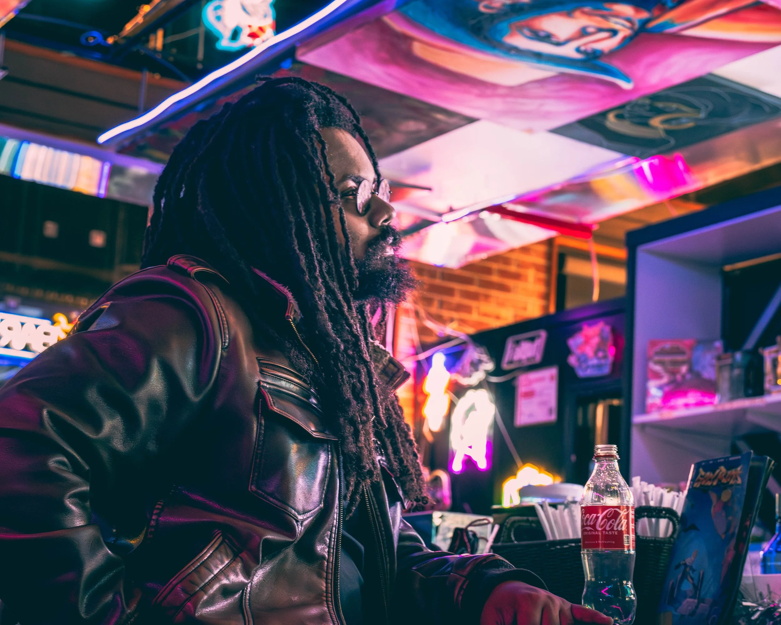 A man with dreadlocks and glasses is sitting at a table in a neon-lit bar or arcade, with a Coca-Cola bottle and other bottles on the table. The ceiling and walls are decorated with colorful neon signs.