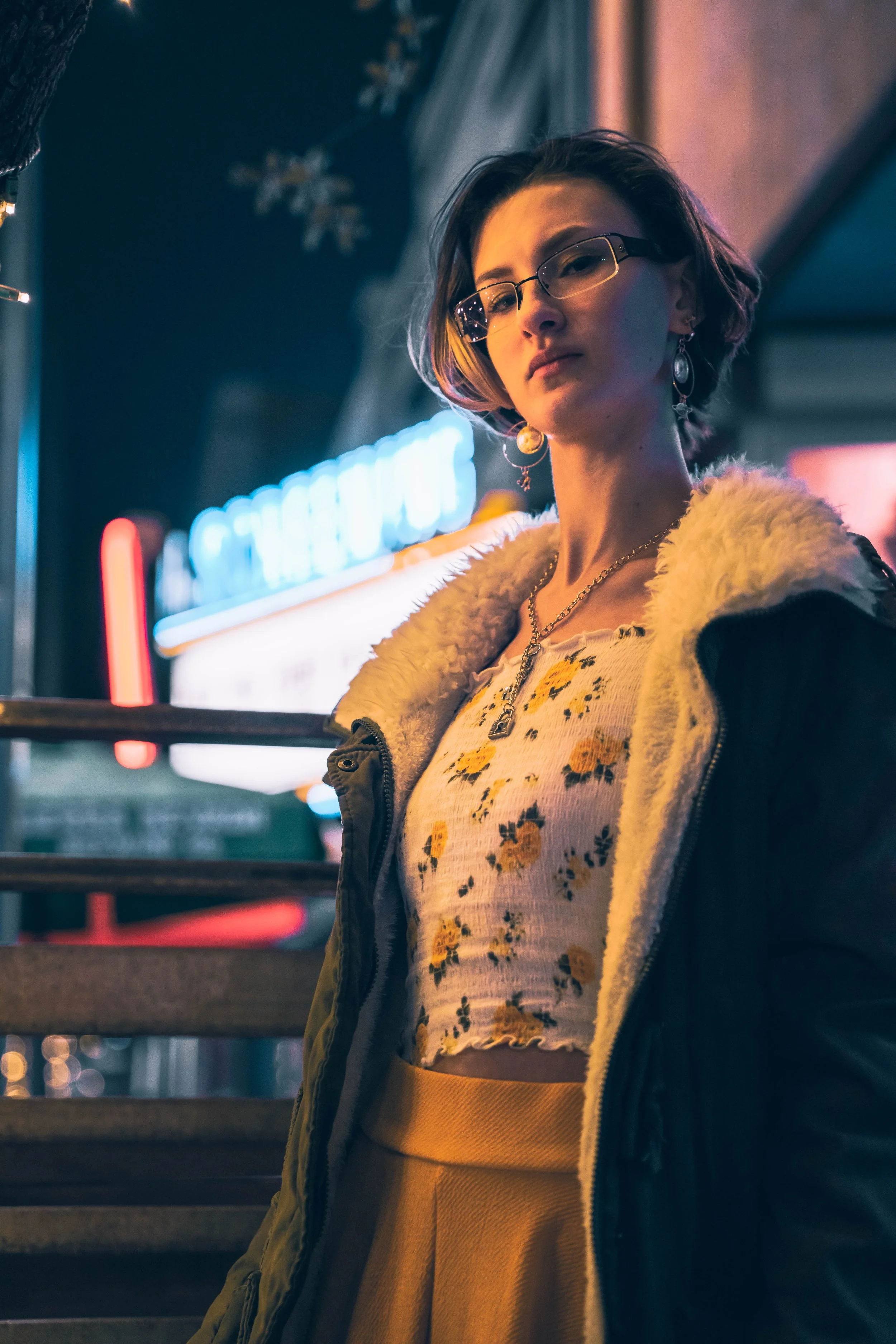 A young woman with short hair wearing glasses, earrings, a yellow floral shirt, and a dark jacket with a fur collar, standing outside at night near neon signs and stairs.
