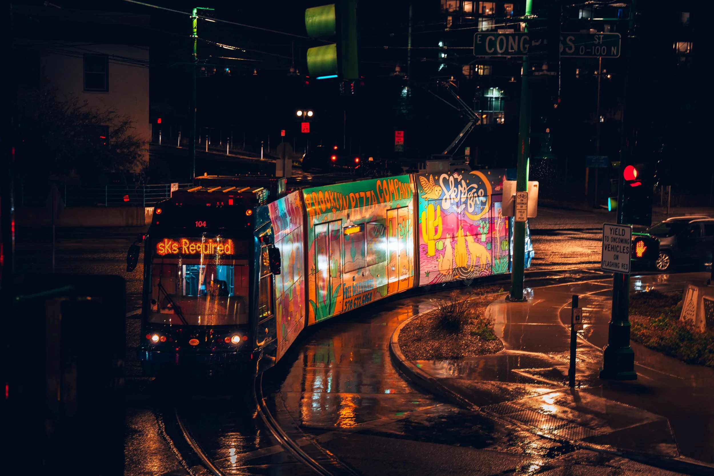 Nighttime city street scene showing a colorful bus with bright artwork, wet streets reflecting lights, traffic signals, and street signs.