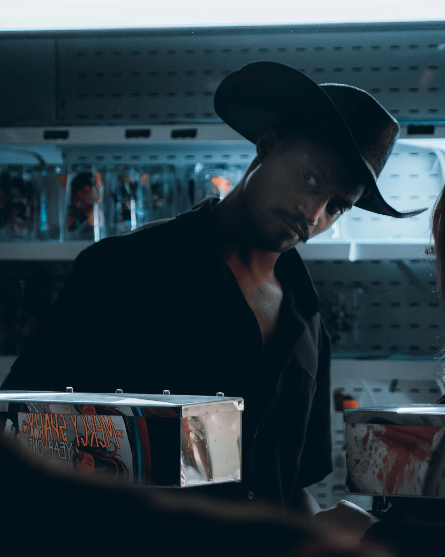 A man wearing a black cowboy hat reflects in a store mirror behind a pizza box with a promotional poster, with shelves of candies or snacks in the background.