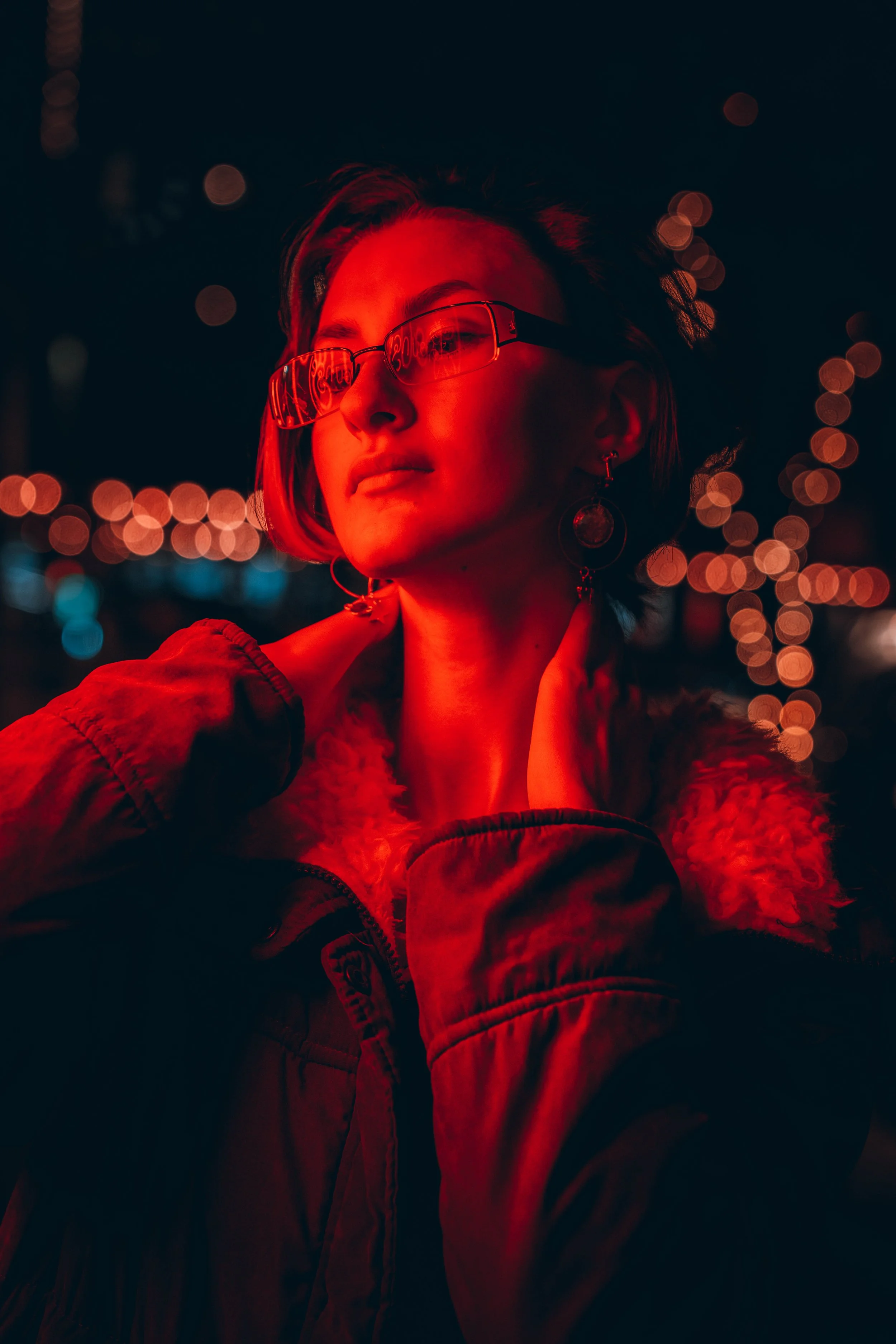 Portrait of a young woman with short dark hair and glasses, illuminated by red neon lighting, with blurred lights in the background.