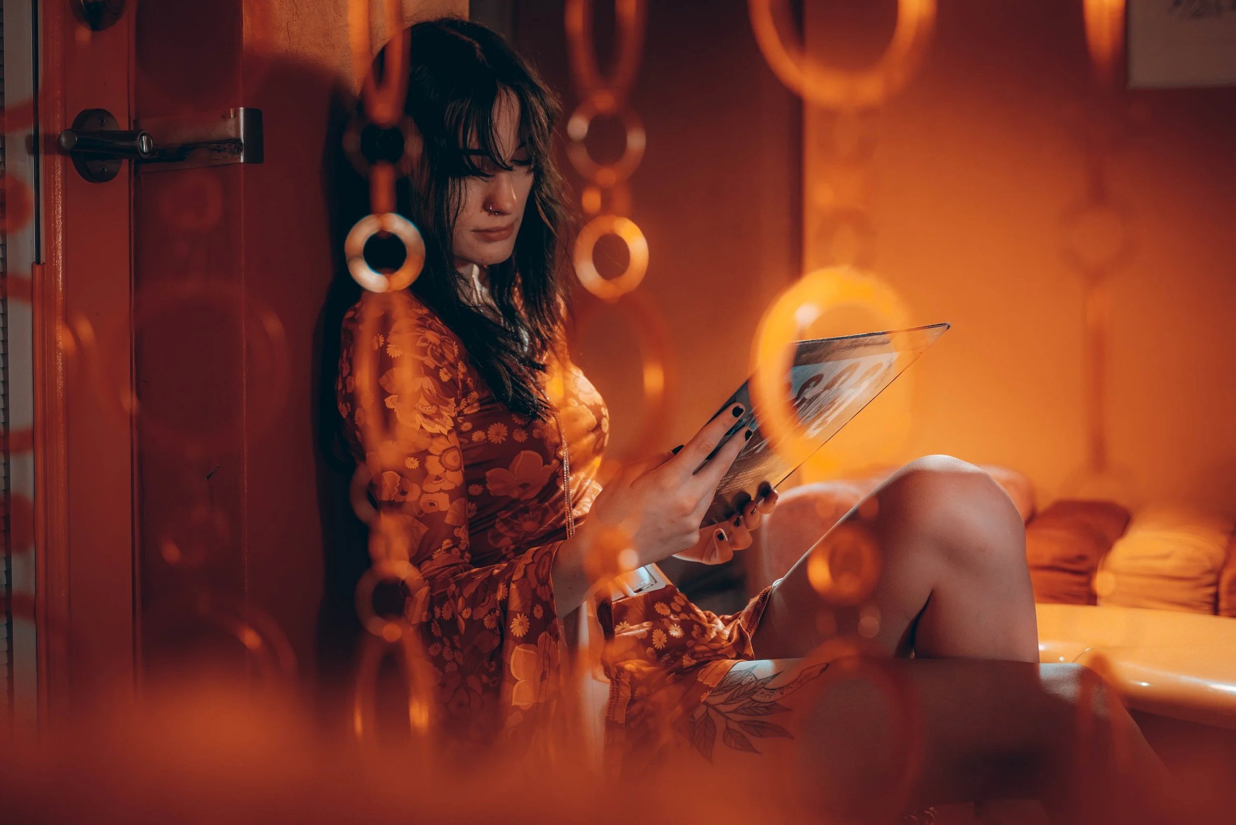 A young woman with dark hair, glasses, and a nose ring sitting on a bed against a wooden wall, reading a magazine or book in a warm, orange-lit room.