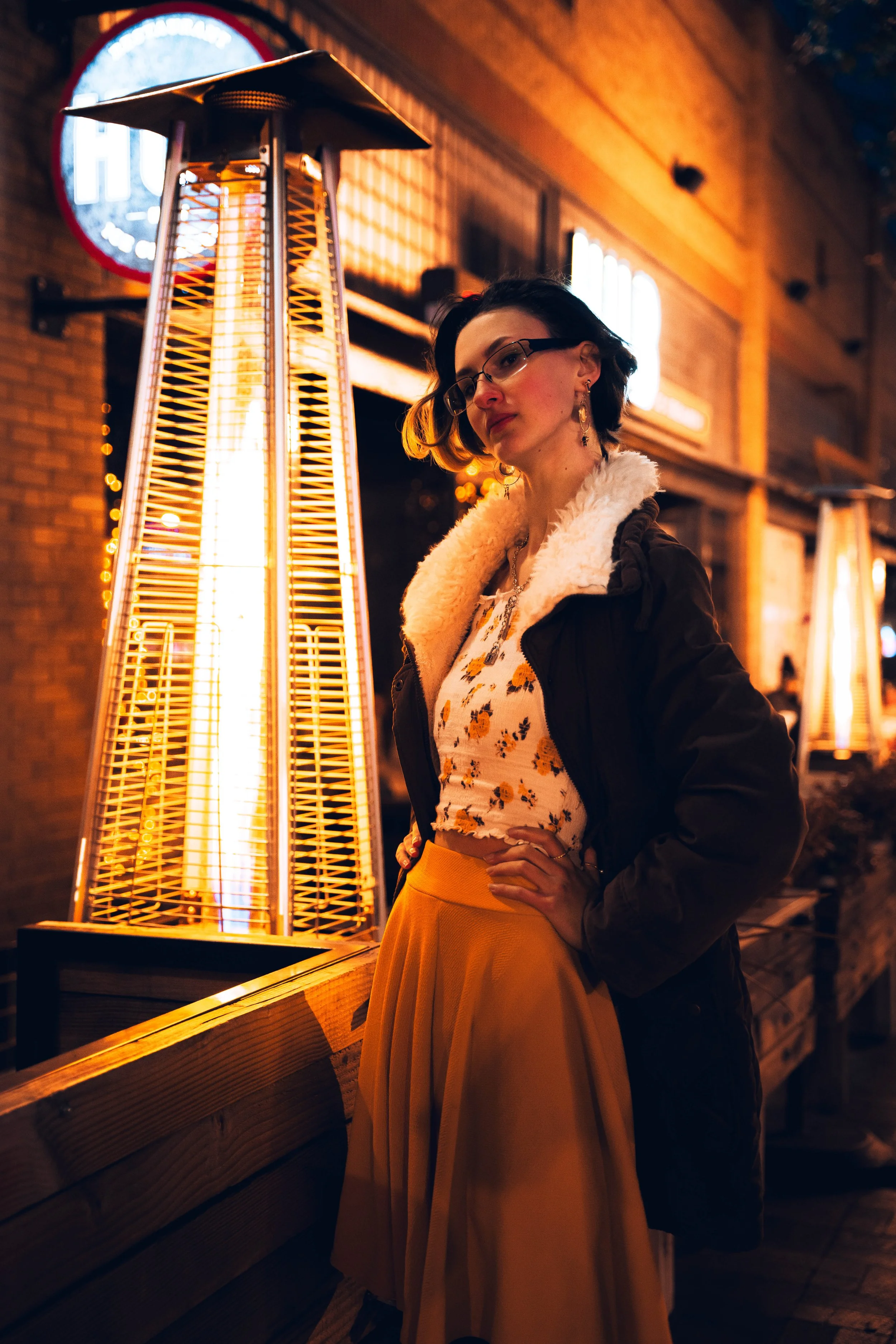 A woman wearing glasses, a floral top, and a black jacket with a shearling collar, standing next to a large outdoor patio heater at night.