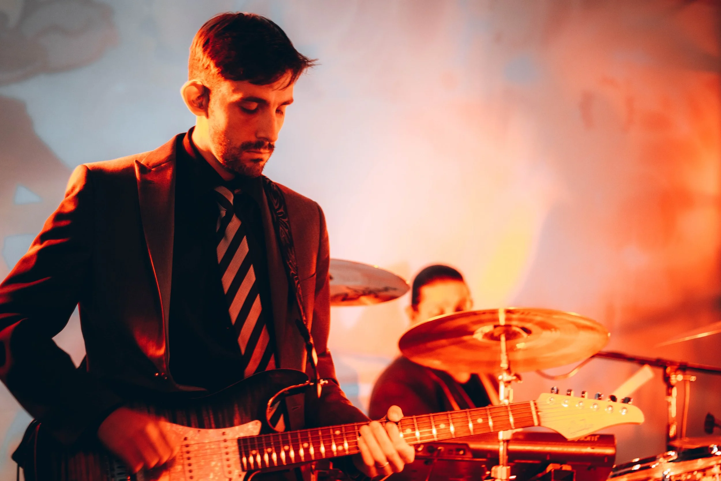 A man playing electric guitar during a live concert with orange and red stage lighting, accompanied by a drummer in the background.