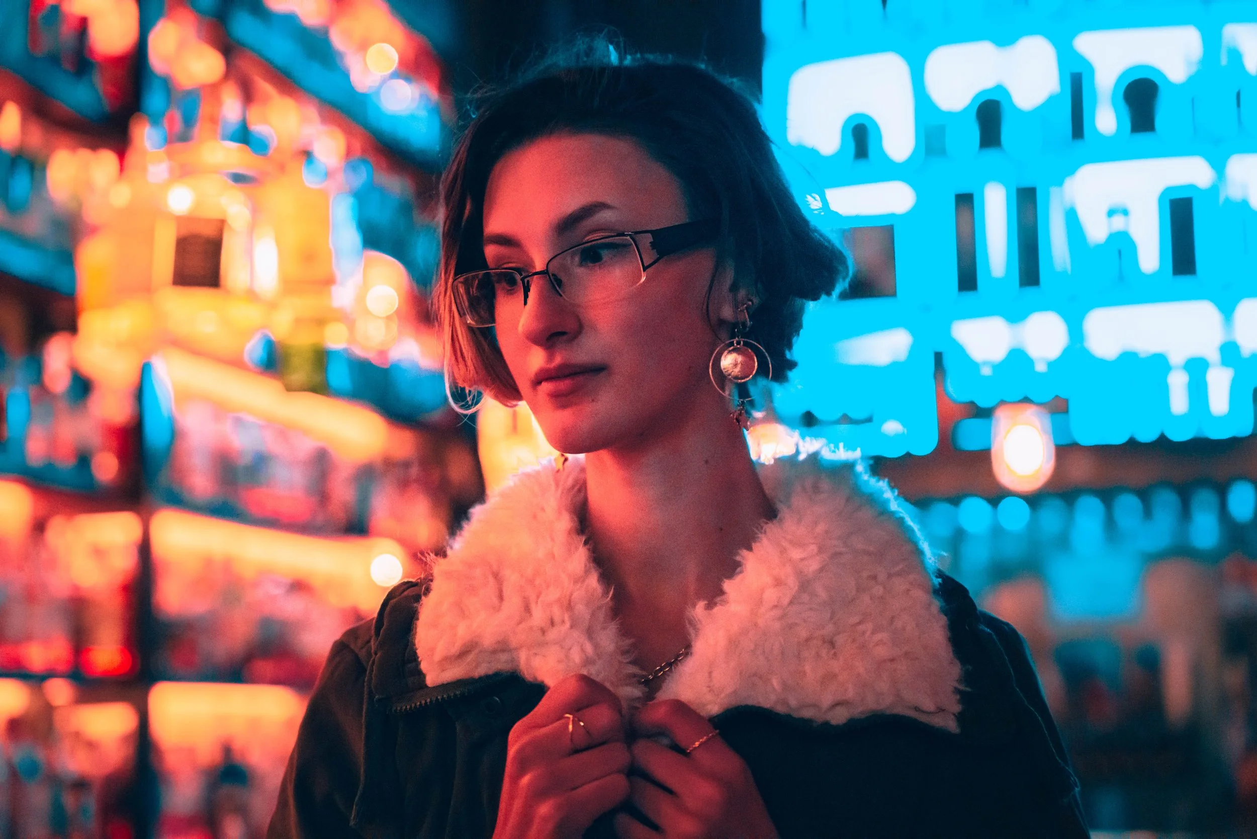 A young woman with short dark hair, glasses, and earrings is standing at night surrounded by colorful neon lights on a city street. She is wearing a fuzzy white coat and a black jacket and appears to be adjusting her clothing.