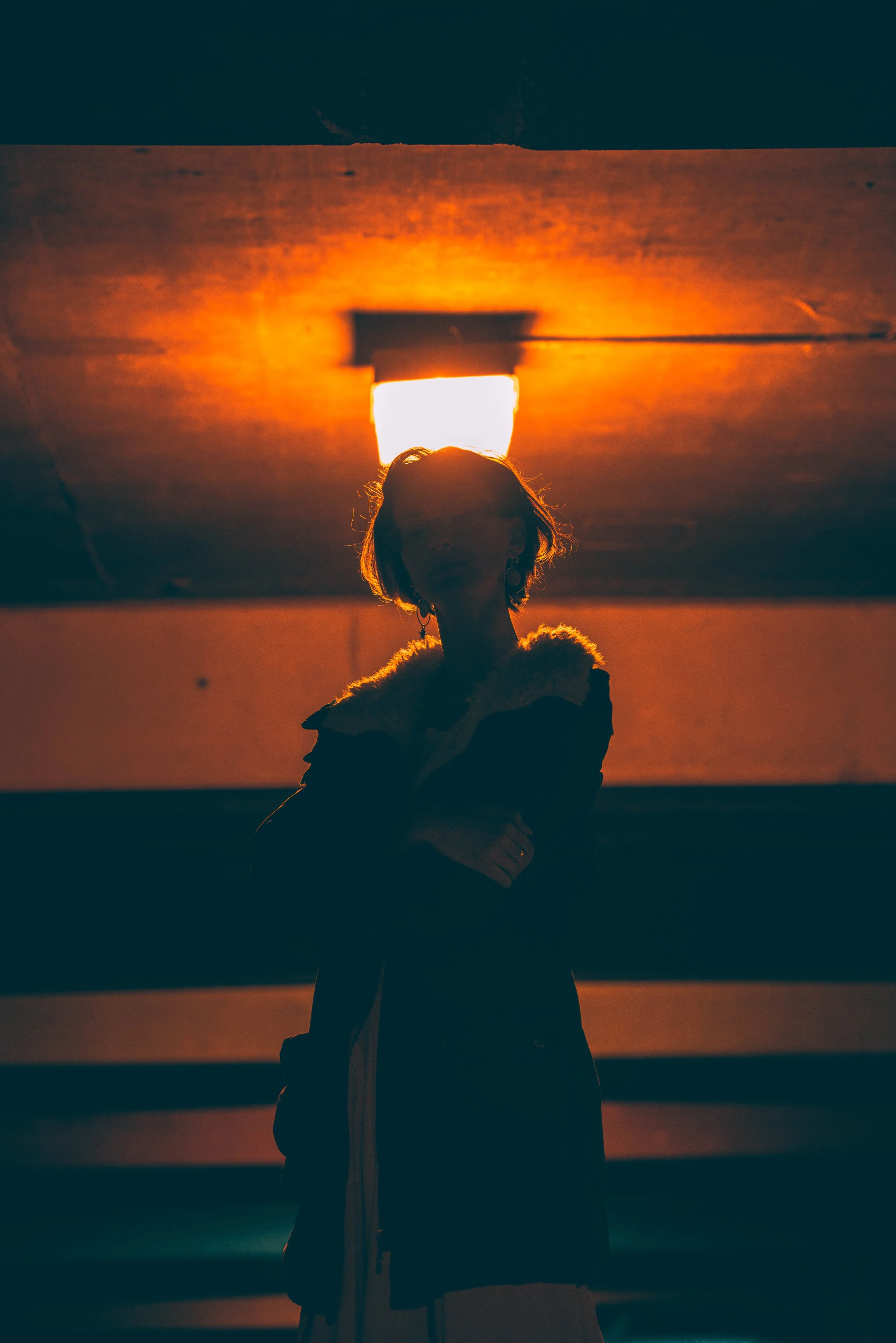 Person standing in front of a bright light, facing away. The image is dark, with the person's hair and clothing visible, illuminated slightly by the light.