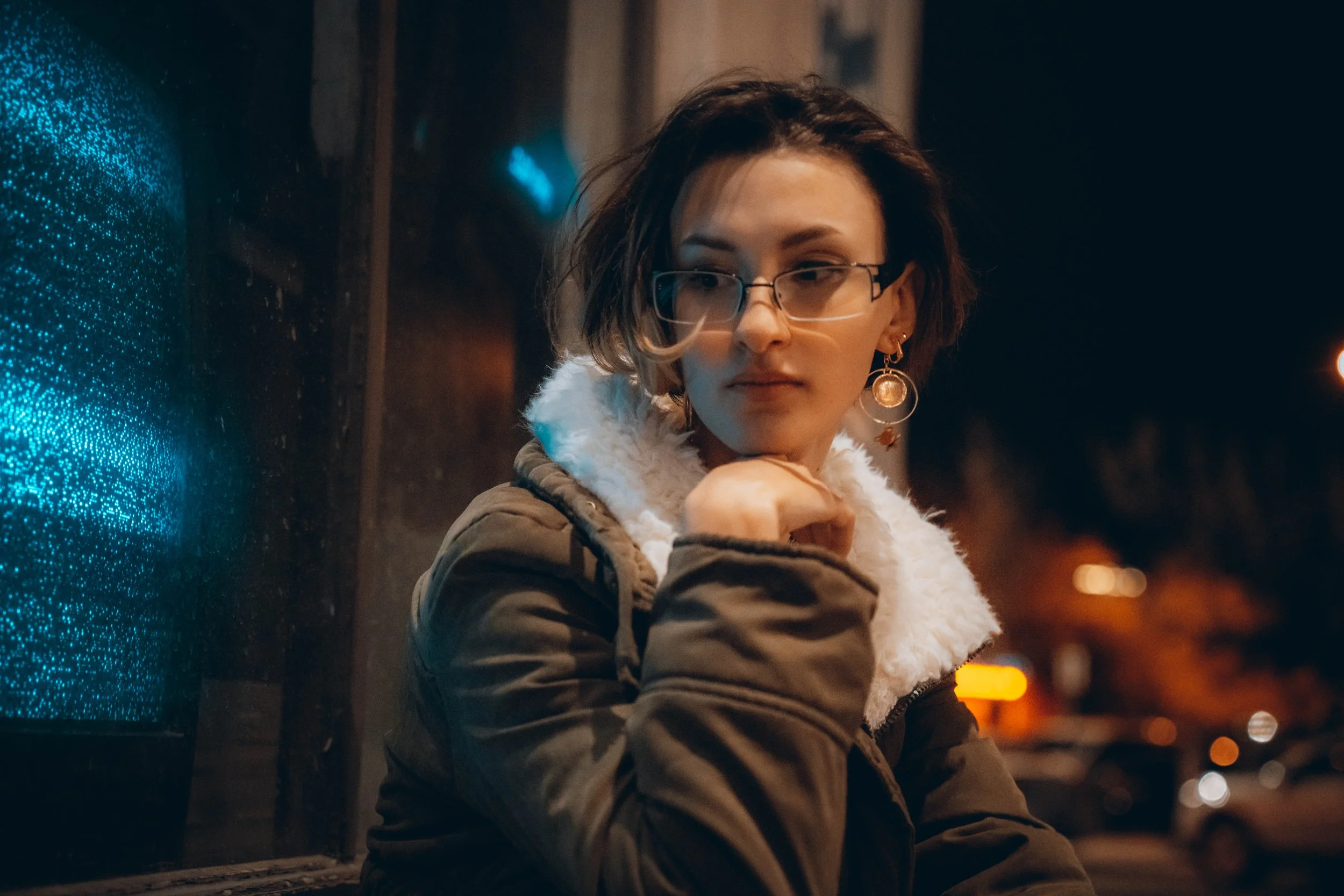 A woman with short dark hair, glasses, and earrings, wearing a hoodie with a white fuzzy lining, is leaning on a metal rail at night. She appears to be looking at something or someone, with city lights blurred in the background.