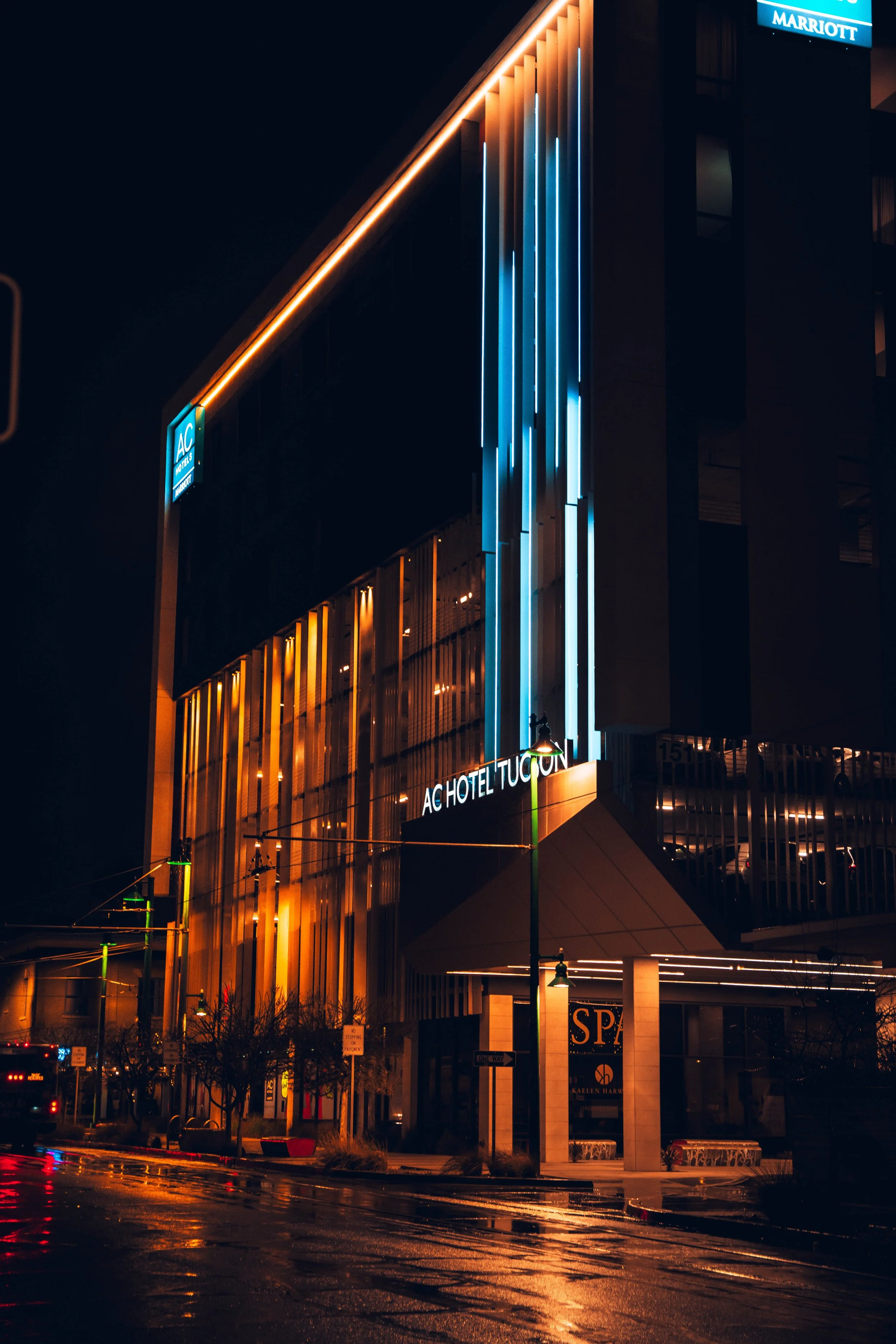 Night view of a modern hotel building with neon blue and warm yellow lighting, wet street reflections, and a sign indicating 'AC HOTEL TUCON' and 'SPA.'