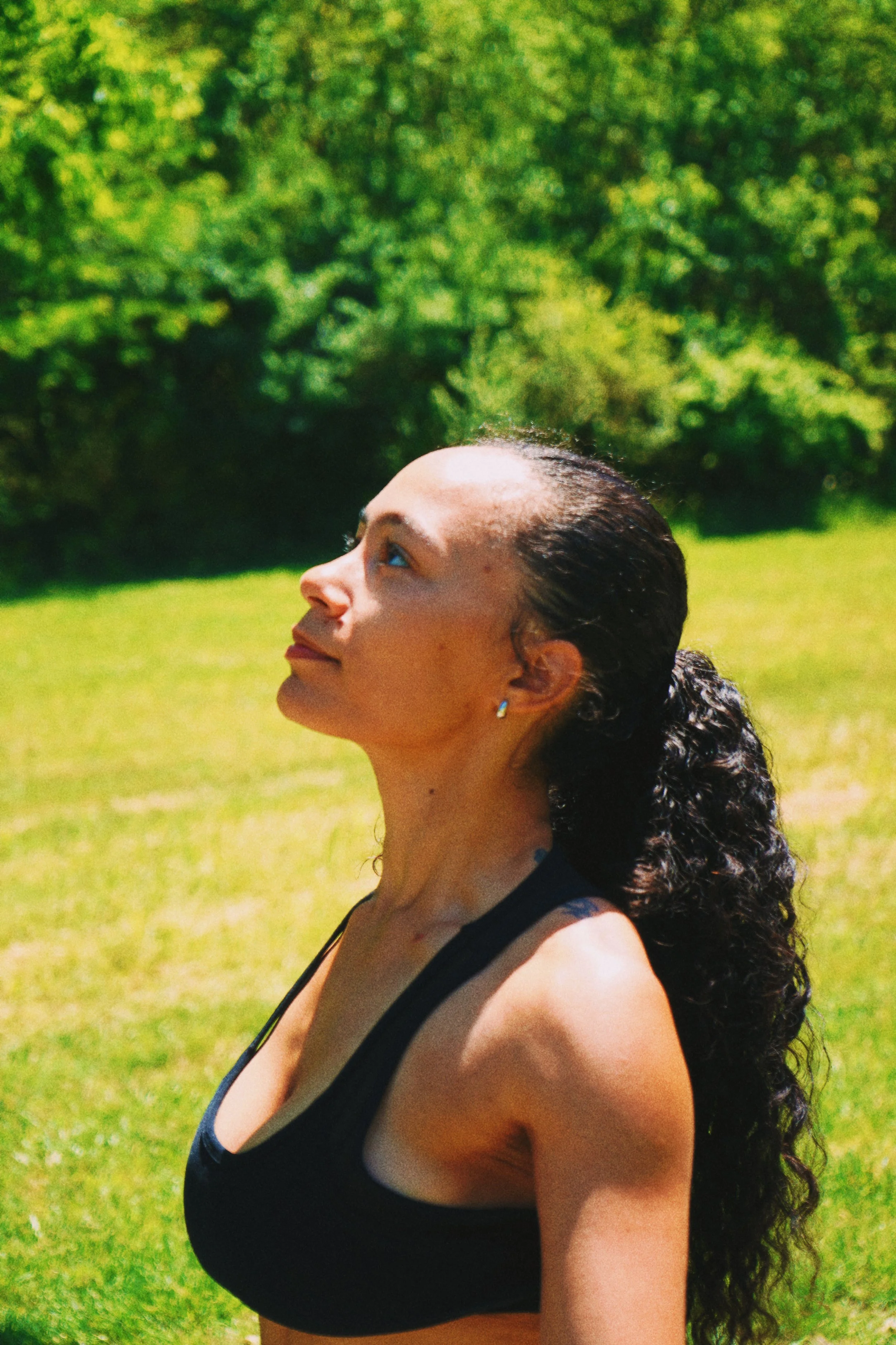 Profile of a woman with dark, curly hair tied back, wearing a black sports bra, standing outdoors on green grass with trees in the background.