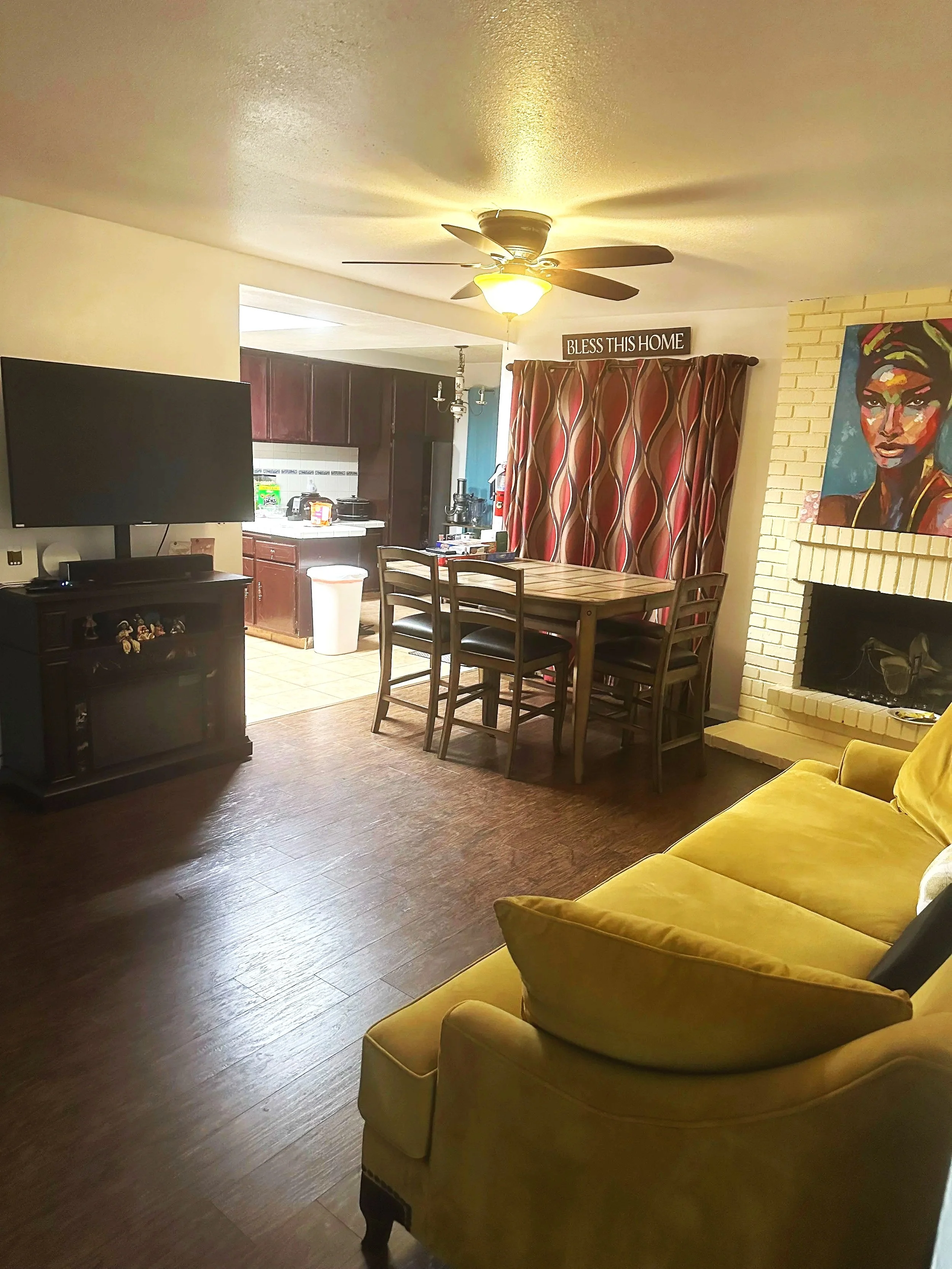 A living room with a yellow sofa, a wall-mounted flat-screen TV, wooden dining table with four chairs, kitchen in the background, a sign saying 'Bless This Home', colorful curtain over a doorway, colorful artwork of a woman's face on a brick fireplace, ceiling fan with light, and hardwood flooring.