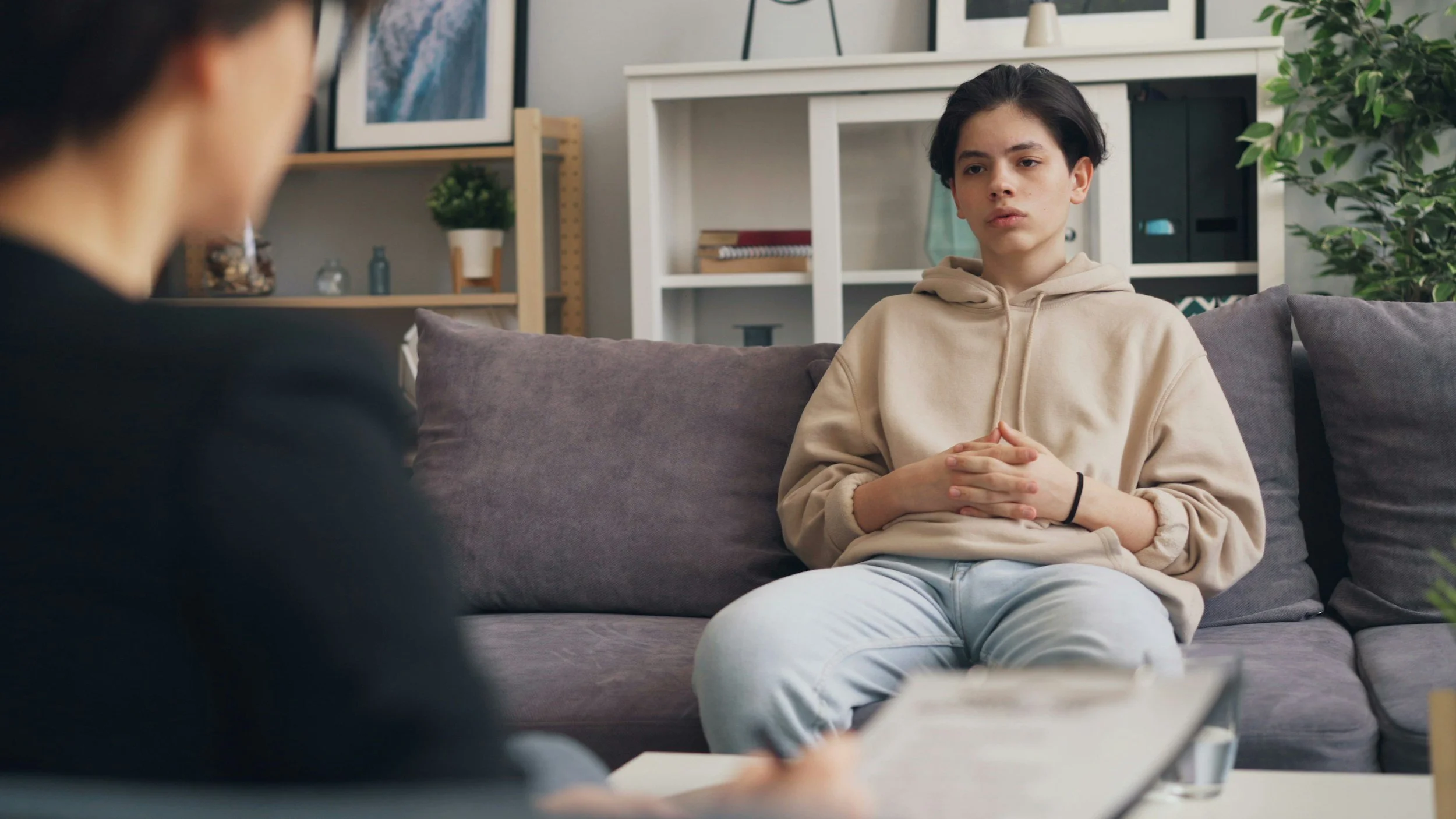 Young woman in beige hoodie sitting on a gray couch with her hands clasped, talking to a person whose face is not visible, in a modern living room with bookshelves, plants, and framed pictures.