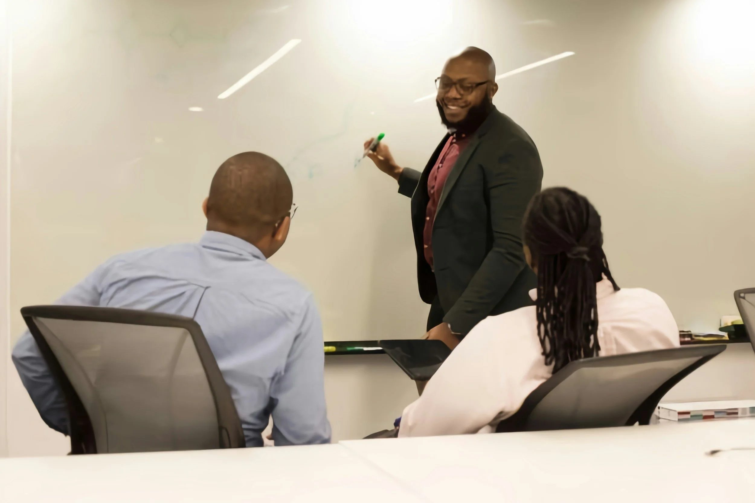 Man in a black blazer and glasses writing on a whiteboard during a meeting with three people seated at a conference table.