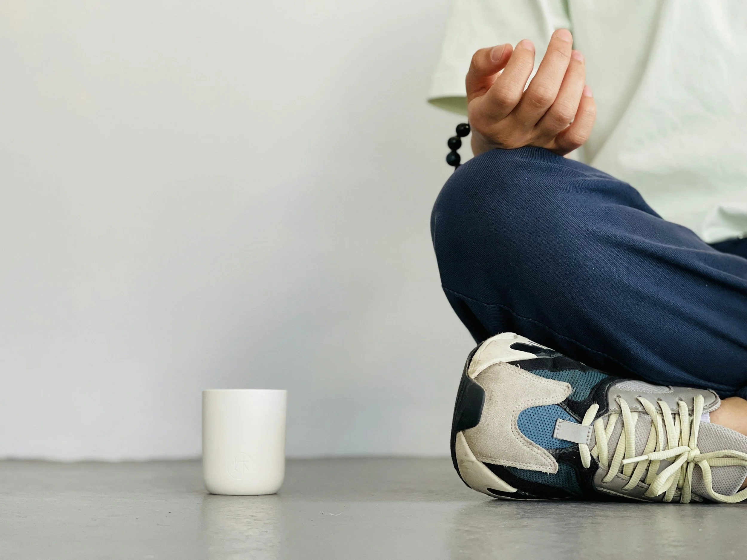A person squatting on one leg with hands in a prayer position, wearing athletic shoes and a bracelet, next to a white ceramic cup.