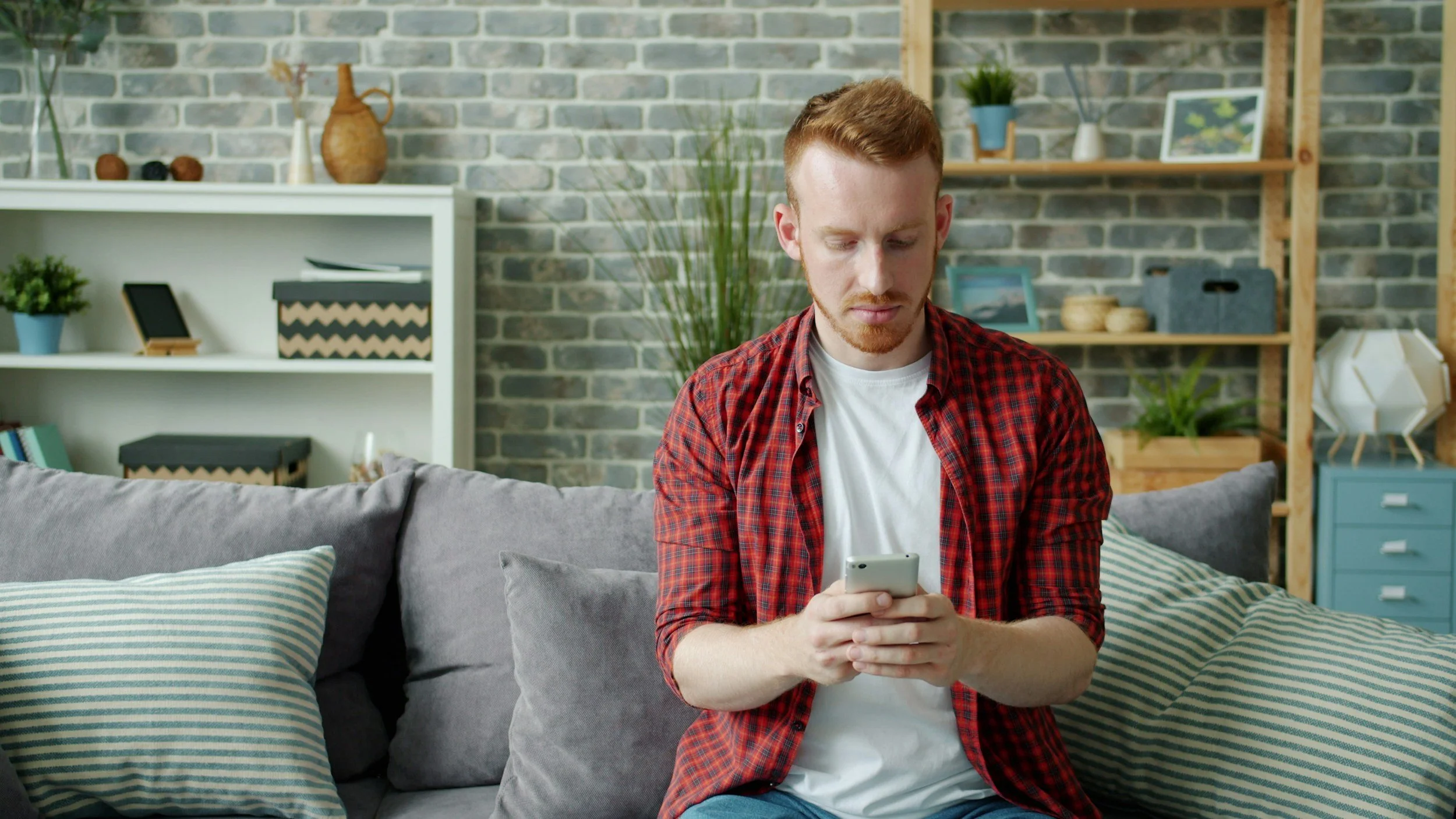 A young man with red hair and a beard, wearing a red plaid shirt over a white T-shirt, is sitting on a sofa in a living room, looking at his smartphone.