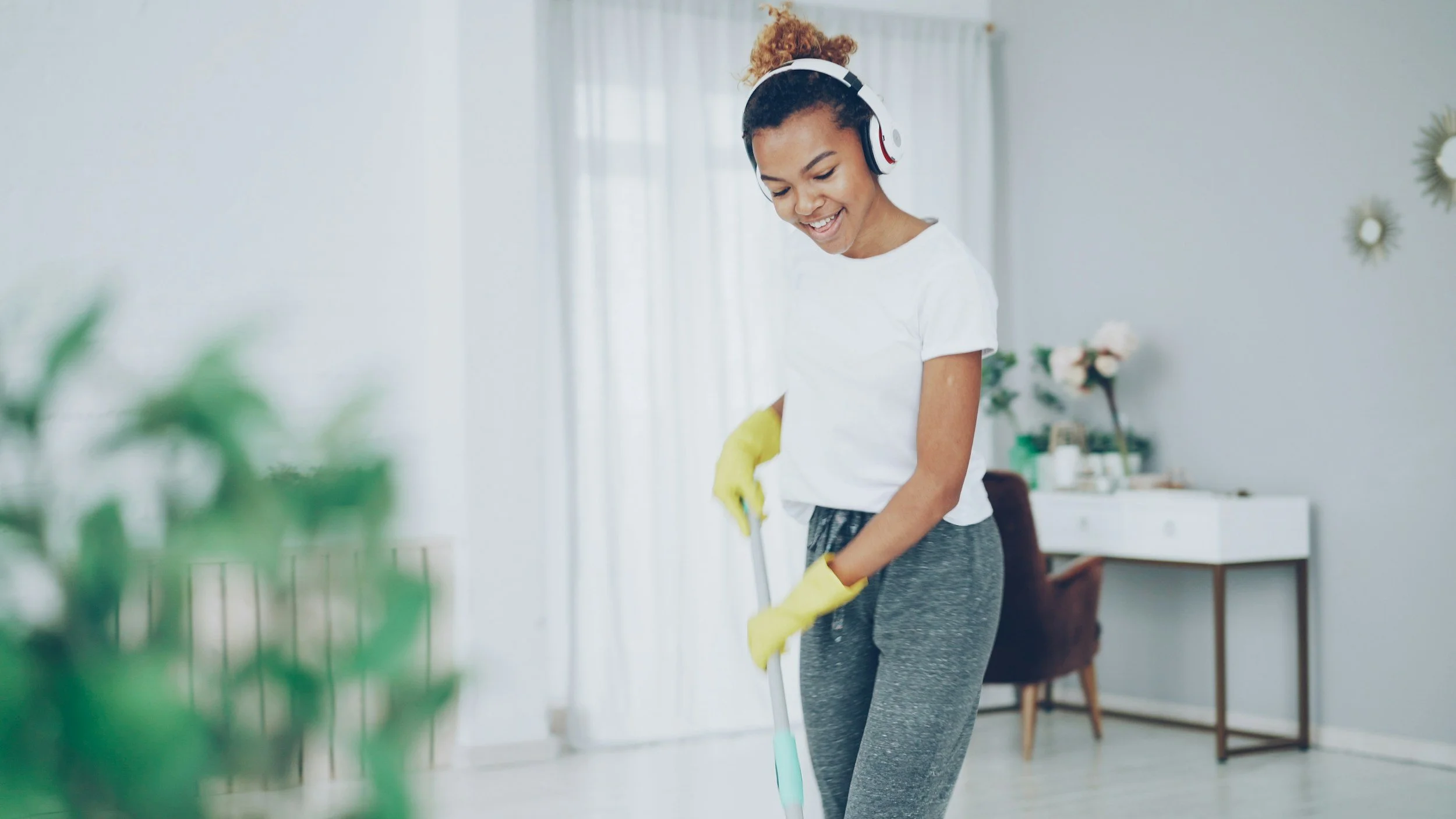 A woman smiling while cleaning the floor with a mop, wearing yellow gloves and headphones in a bright living room.