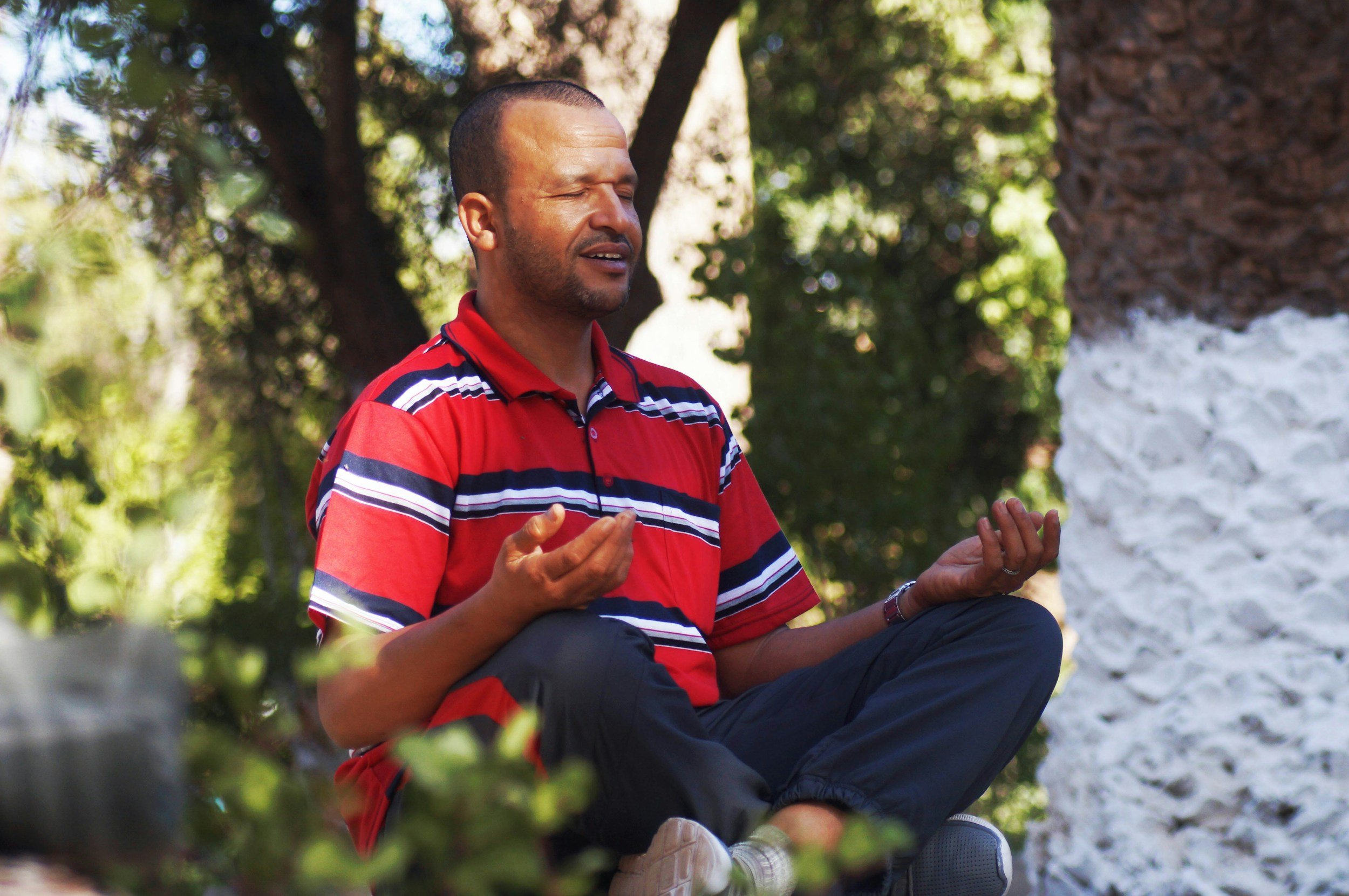 A man sitting outdoors with closed eyes, wearing a red striped polo shirt, appearing to be meditating or praying, surrounded by trees and leaves.