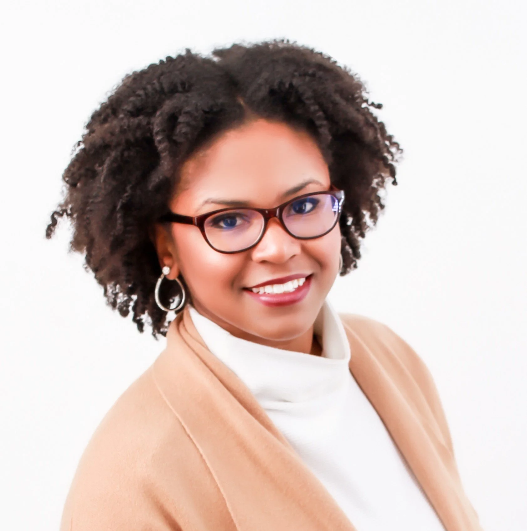 Close-up of a young woman with curly hair wearing glasses, smiling, with a beige blazer and white turtleneck, against a plain white background.