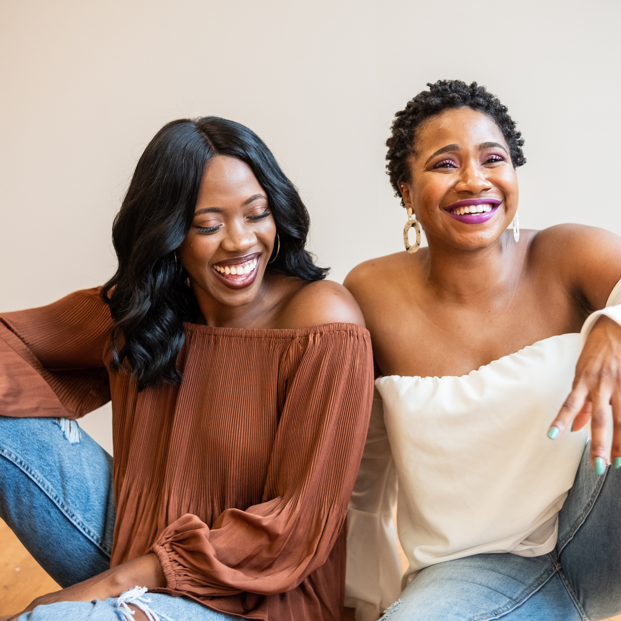 Two women smiling and leaning close together, one with long black hair and the other with short curly hair, wearing off-the-shoulder tops and large earrings.