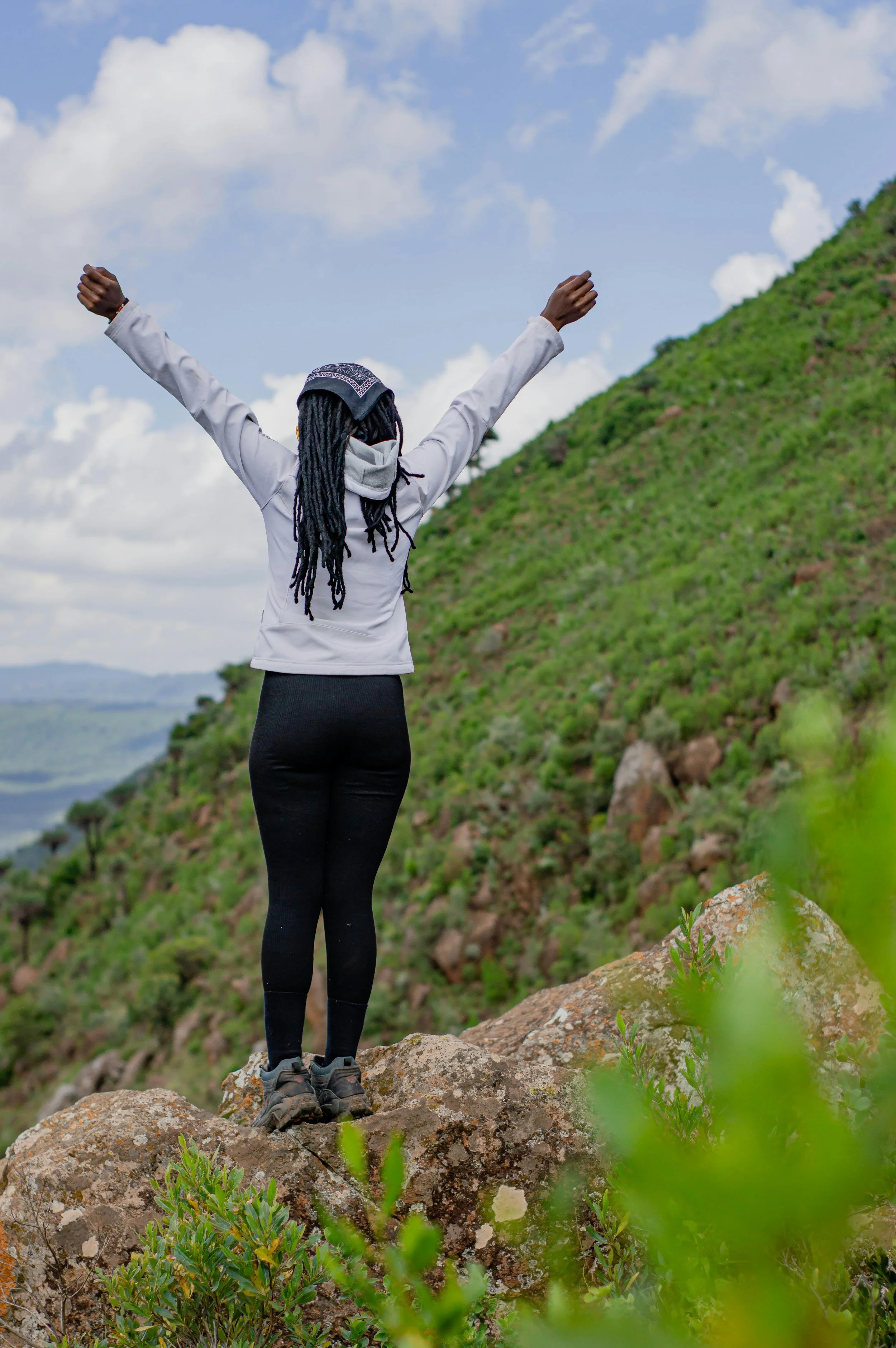 A person with long black dreadlocks, wearing black leggings, gray hiking shoes, a gray jacket, and a bandana on their head, stands on a large rock with arms raised in the air during daytime. They are on a green hillside with rocks and small plants, under a partly cloudy sky.
