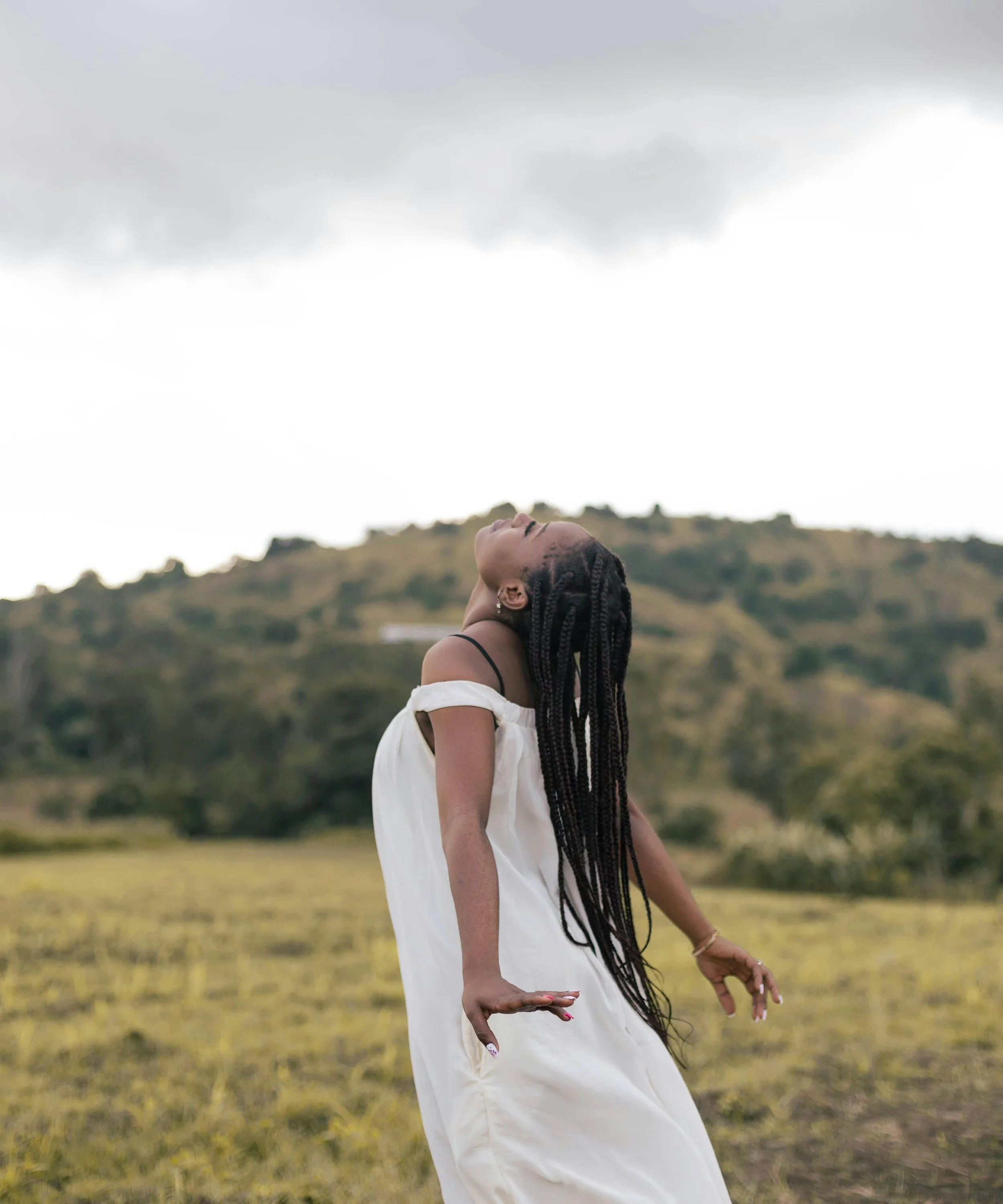 A woman with braids leaning back outdoors in a grassy field with hills, under a cloudy sky.