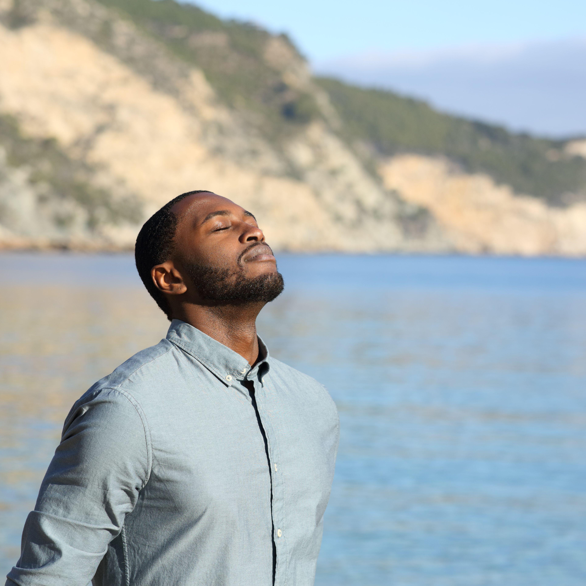 Close-up of an African American man with eyes closed, enjoying the outdoors near water and mountains in the background.