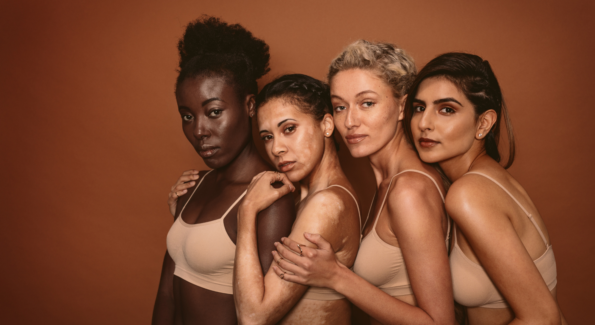 Four diverse women standing close together against a brown background, wearing beige tank tops, with confident expressions.
