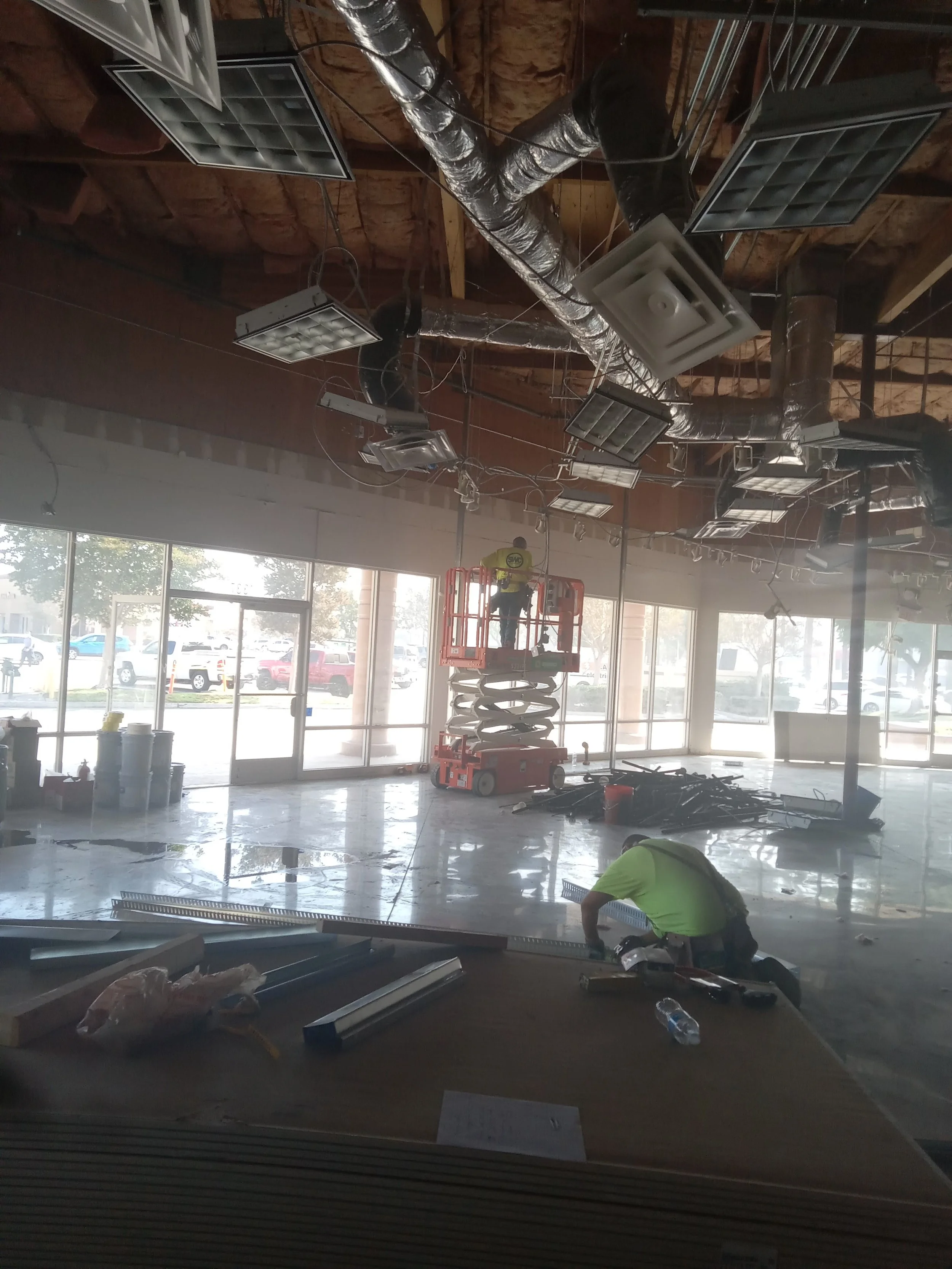 Construction workers installing ceiling fixtures inside a commercial space with large front windows, with tools and materials scattered around.