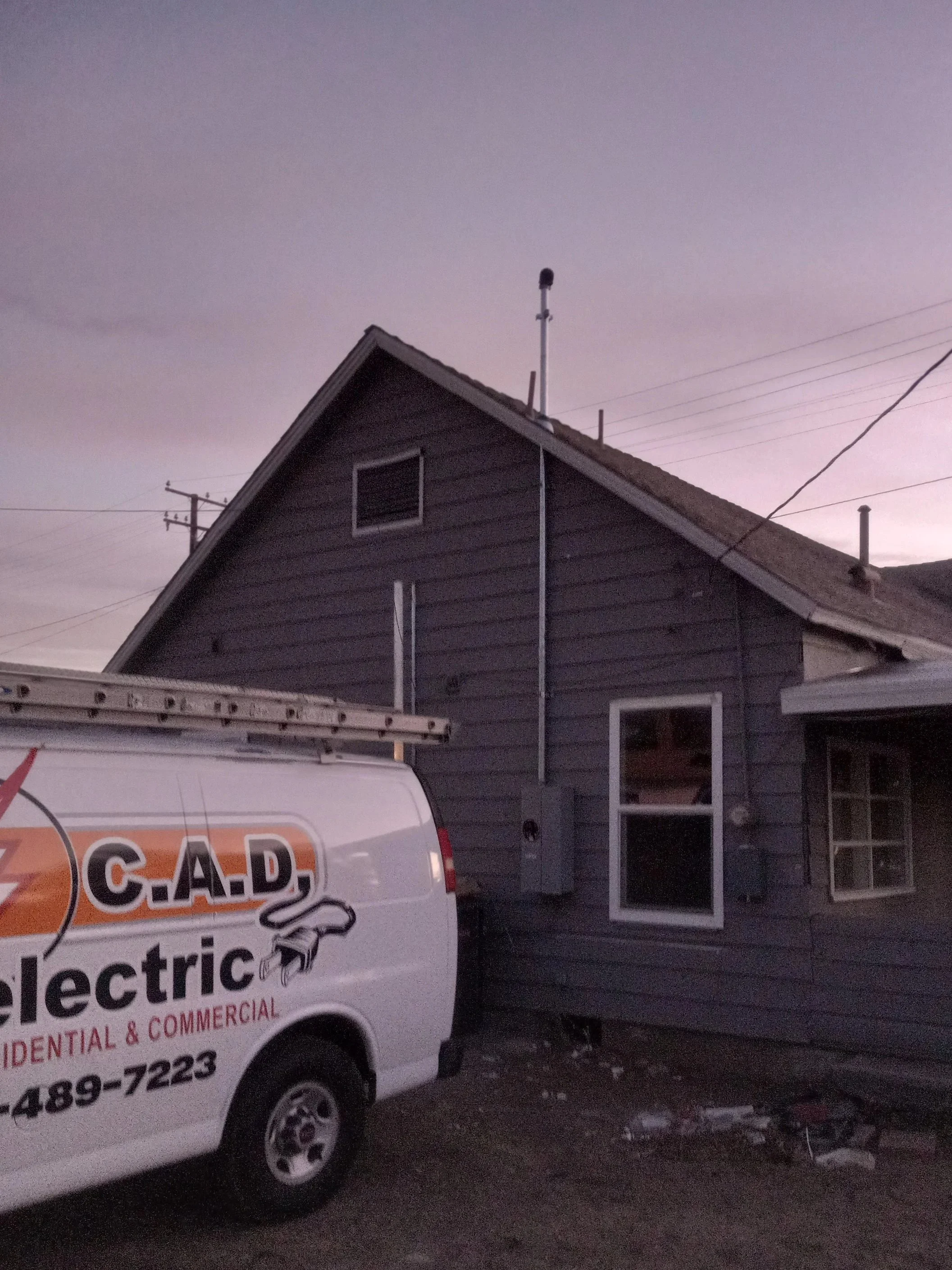 A residential house with gray siding and white trim, with a van labeled 'C.A.D. electric' parked in front. The sky is purple, indicating dawn or dusk.